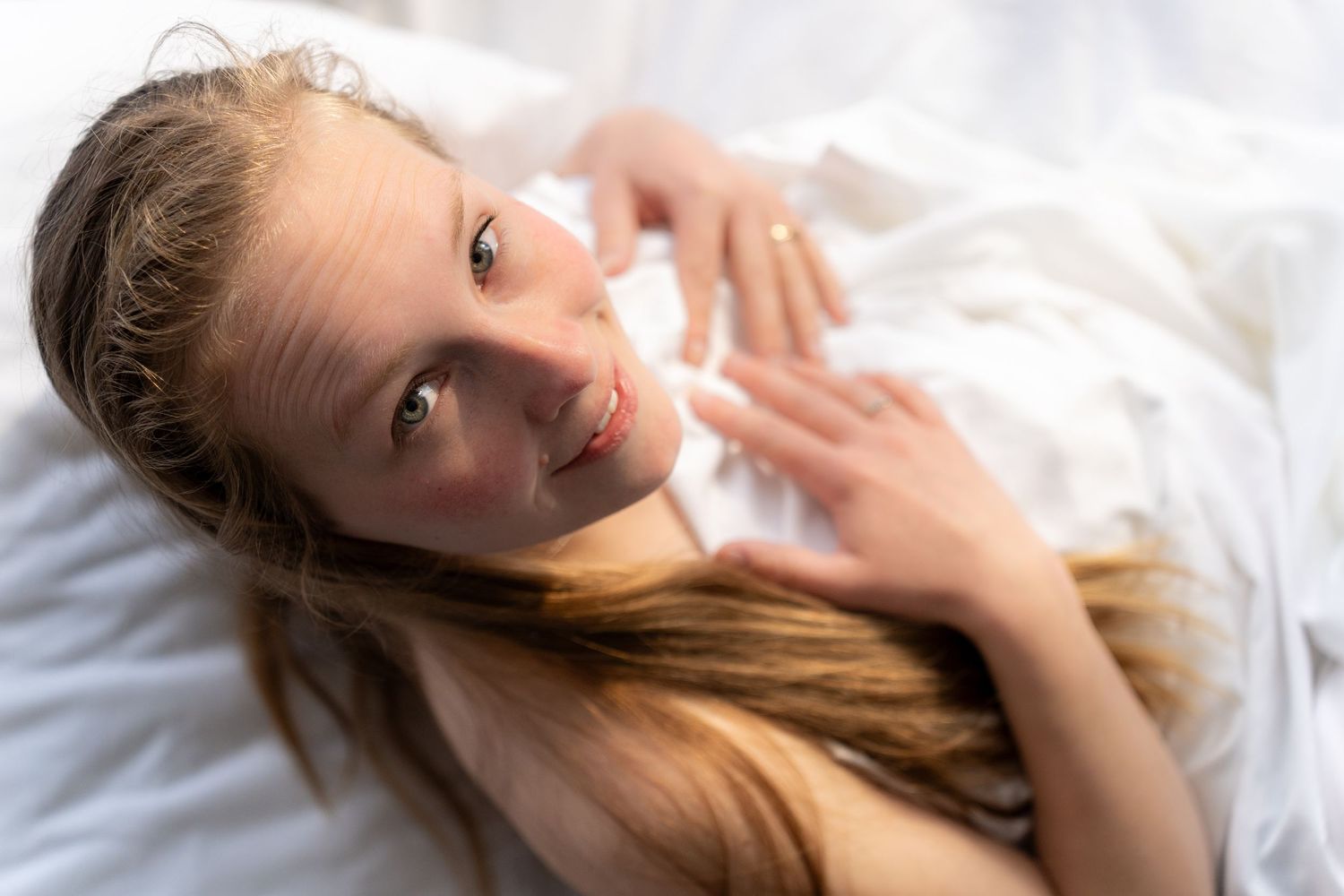 Young girl with long hair lying on white bedding, looking up at the camera with a gentle smile.