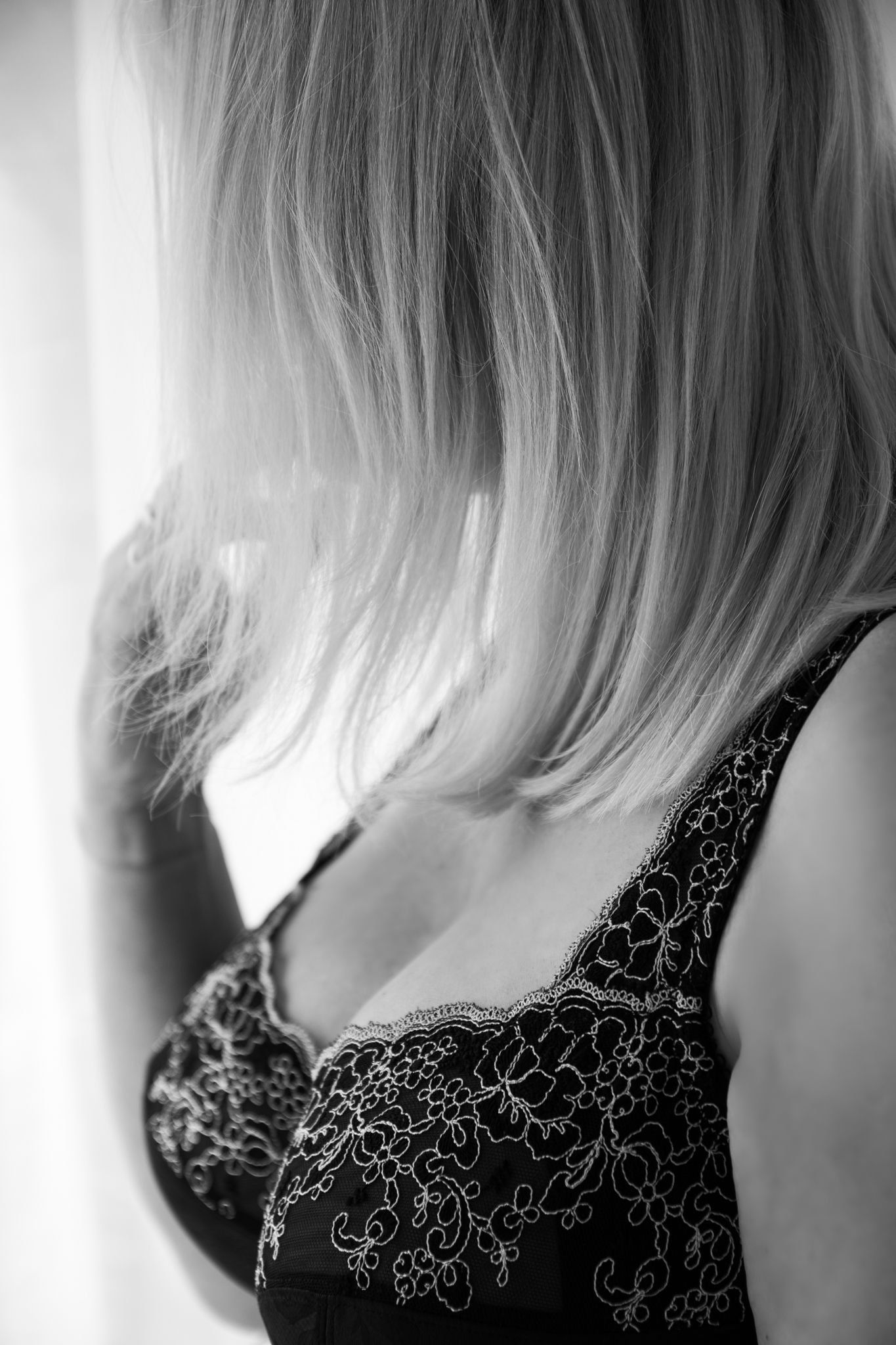 Black and white portrait of a blonde woman in a lace bra standing near a bright window. Boudoir photography Christchurch