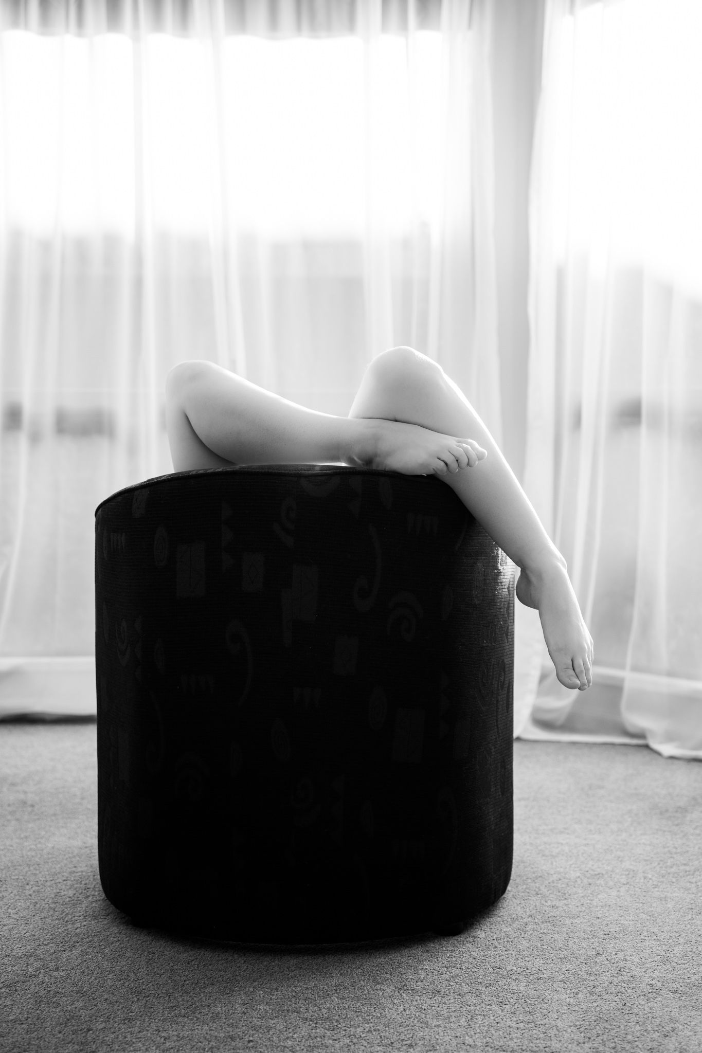 Black and white artistic photo of bare feminine legs resting on a dark round chair near curtains. Boudoir photography Christchurch