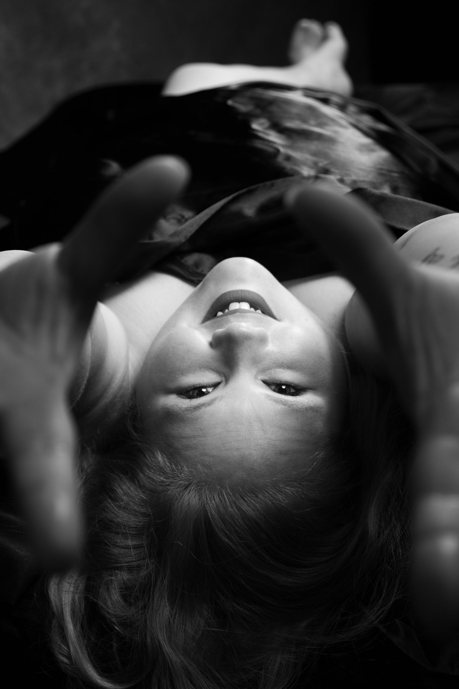 Black and white close-up portrait of a laughing woman lying upside down with hands raised. Boudoir photography Christchurch