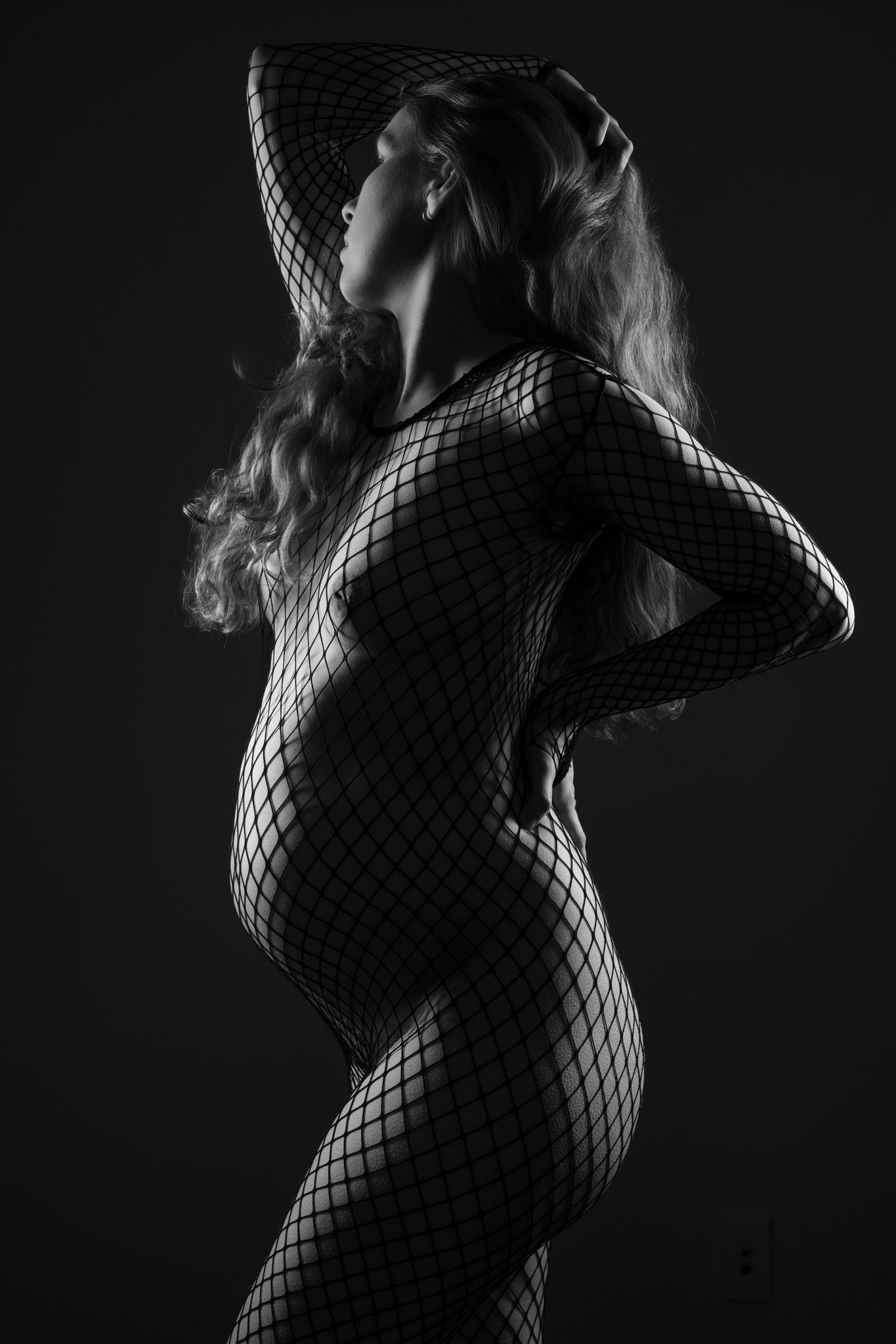 Pregnant woman wearing black fishnet bodysuit posing in dramatic black and white studio portrait. Boudoir photography Christchurch