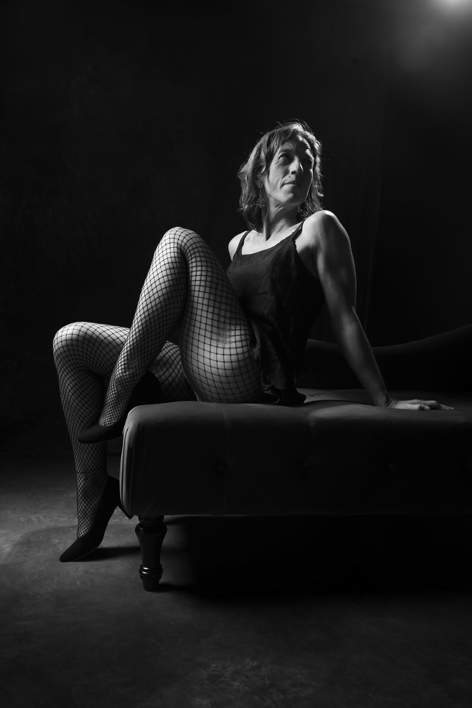 Black and white full-body portrait of a woman in fishnet stockings posing on a dark upholstered bench. Boudoir photography Christchurch
