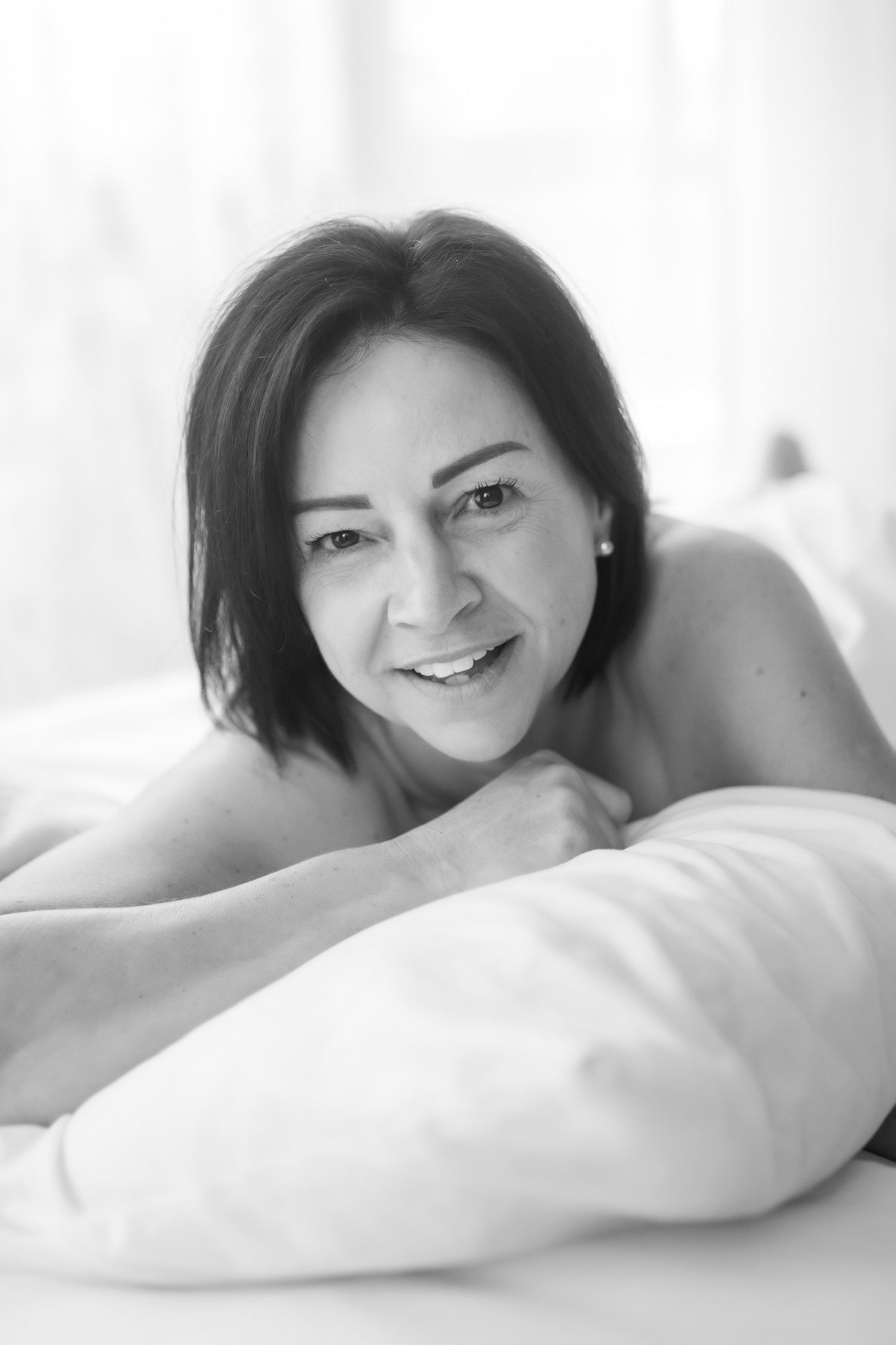 Smiling dark-haired woman resting on white pillow, black and white boudoir portrait photography. Boudoir photography Christchurch