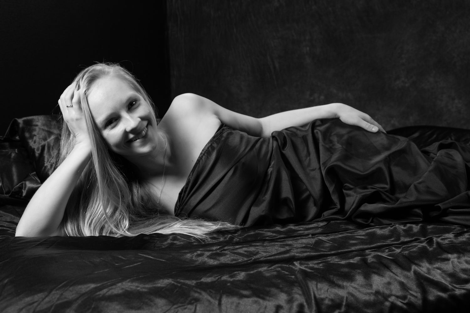 Black and white photo of a smiling blonde woman lying on dark satin bedding in a relaxed pose. Boudoir photography Christchurch