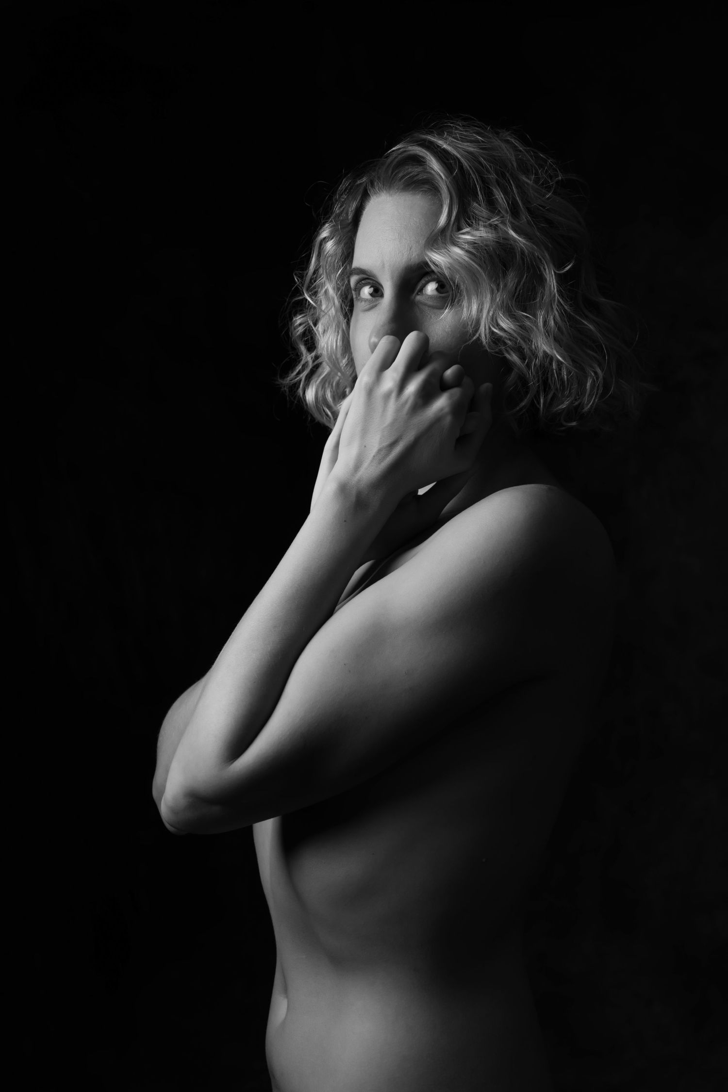 Black and white studio portrait of a woman with curly hair covering her mouth with her hand against dark background. Boudoir photography