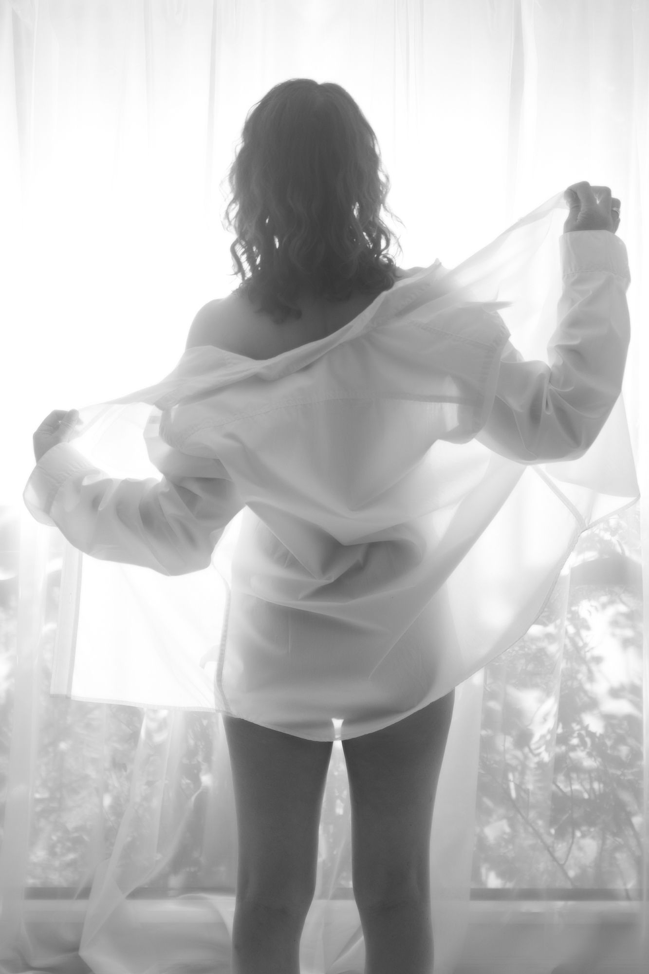 Black and white silhouette of a woman holding sheer white fabric by a bright window. Boudoir photography Christchurch