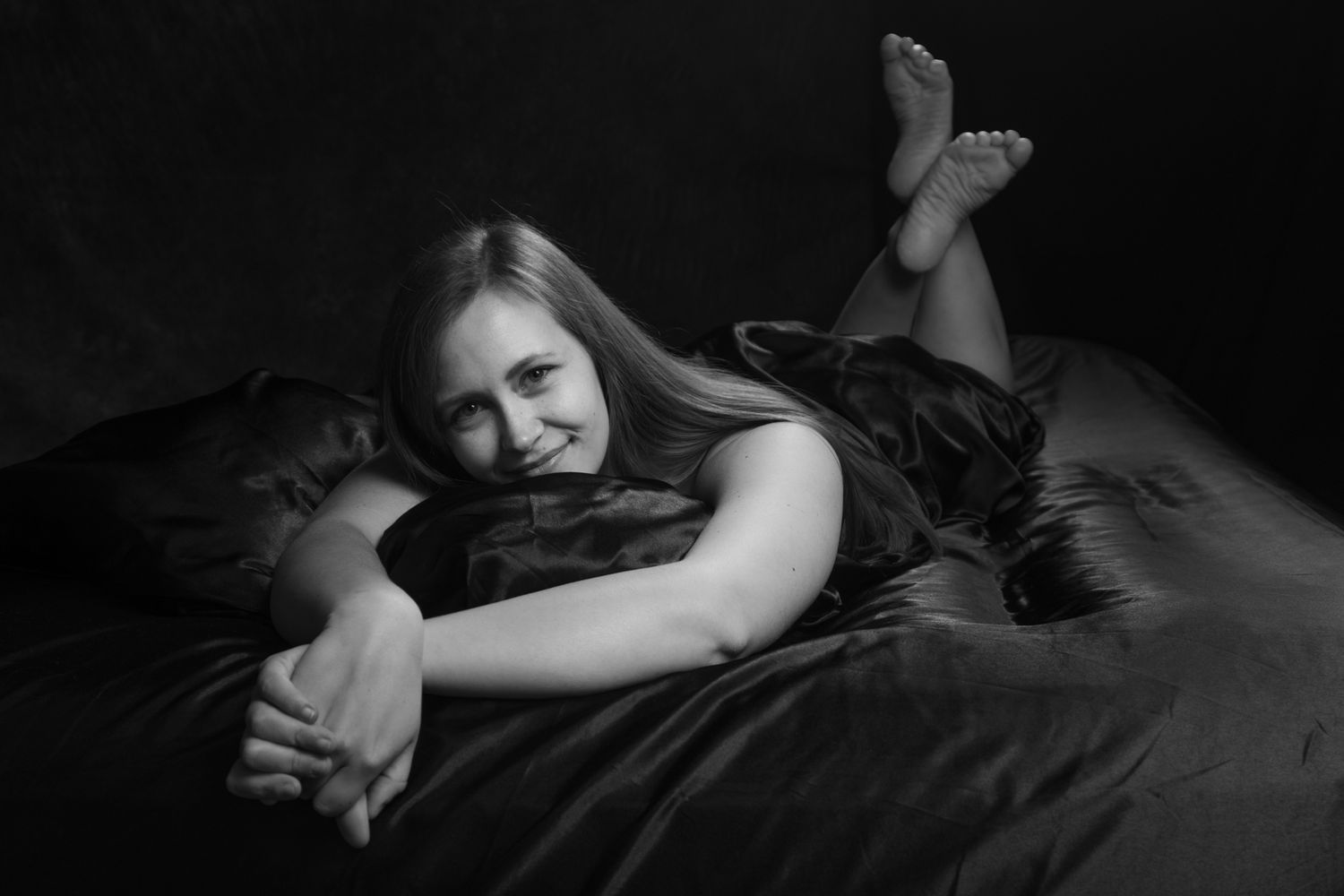 Black and white portrait of a smiling young woman lying on dark bedding with feet raised in a relaxed pose. Boudoir photography Christchurch