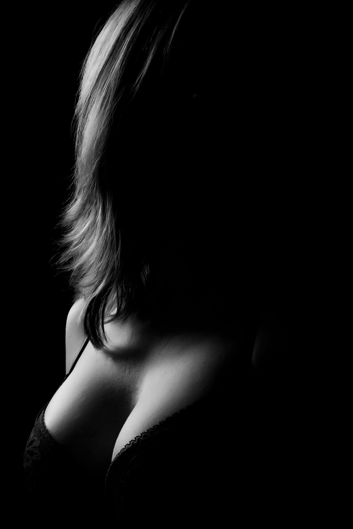 Artistic black and white silhouette of a woman in lingerie, highlighting her shoulder and neckline against a dark background. Boudoir photo