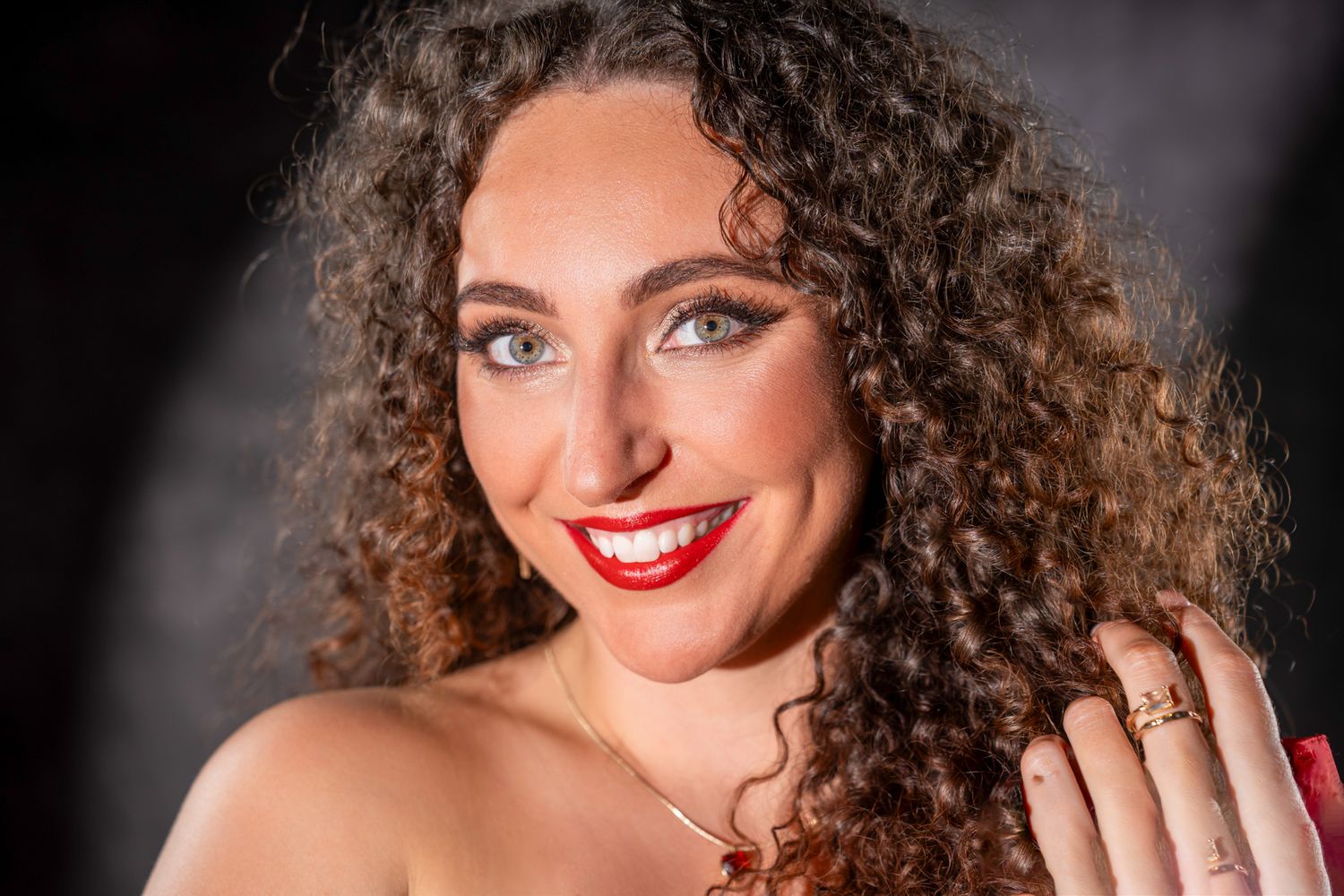 Close-up portrait of a smiling woman with curly hair and red lipstick wearing a gold necklace. Boudoir photography Christchurch portrait