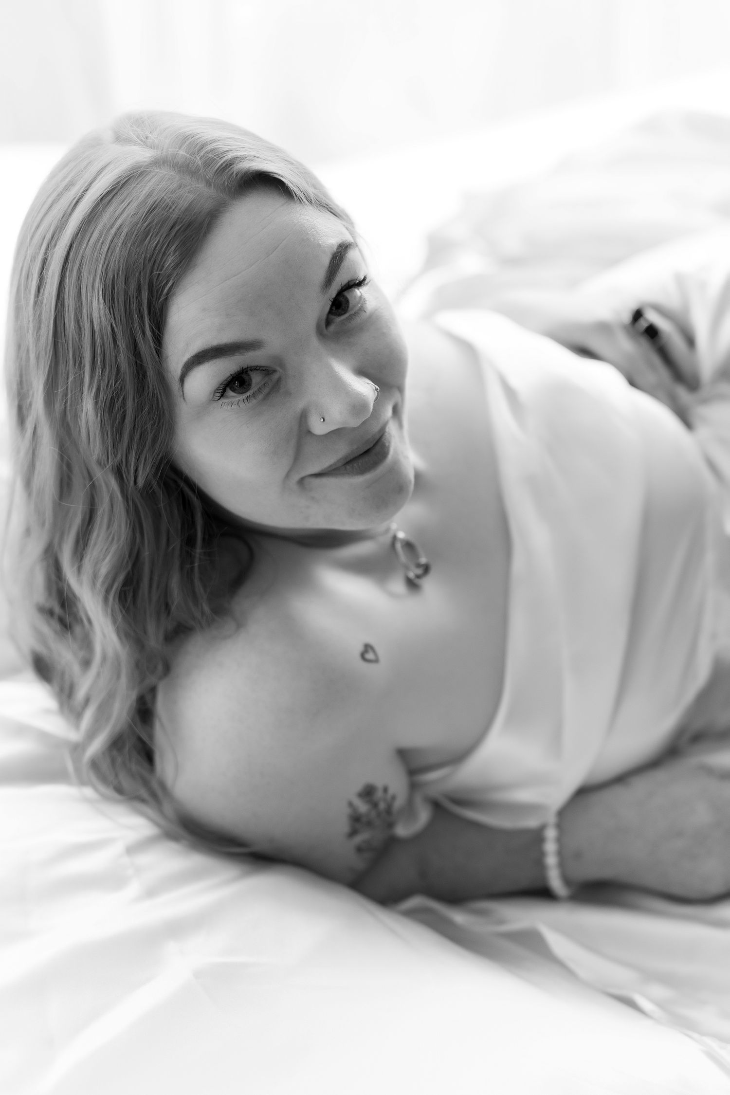 Smiling blonde woman with tattoos lying on white bed, black and white boudoir photography. Boudoir photography Christchurch