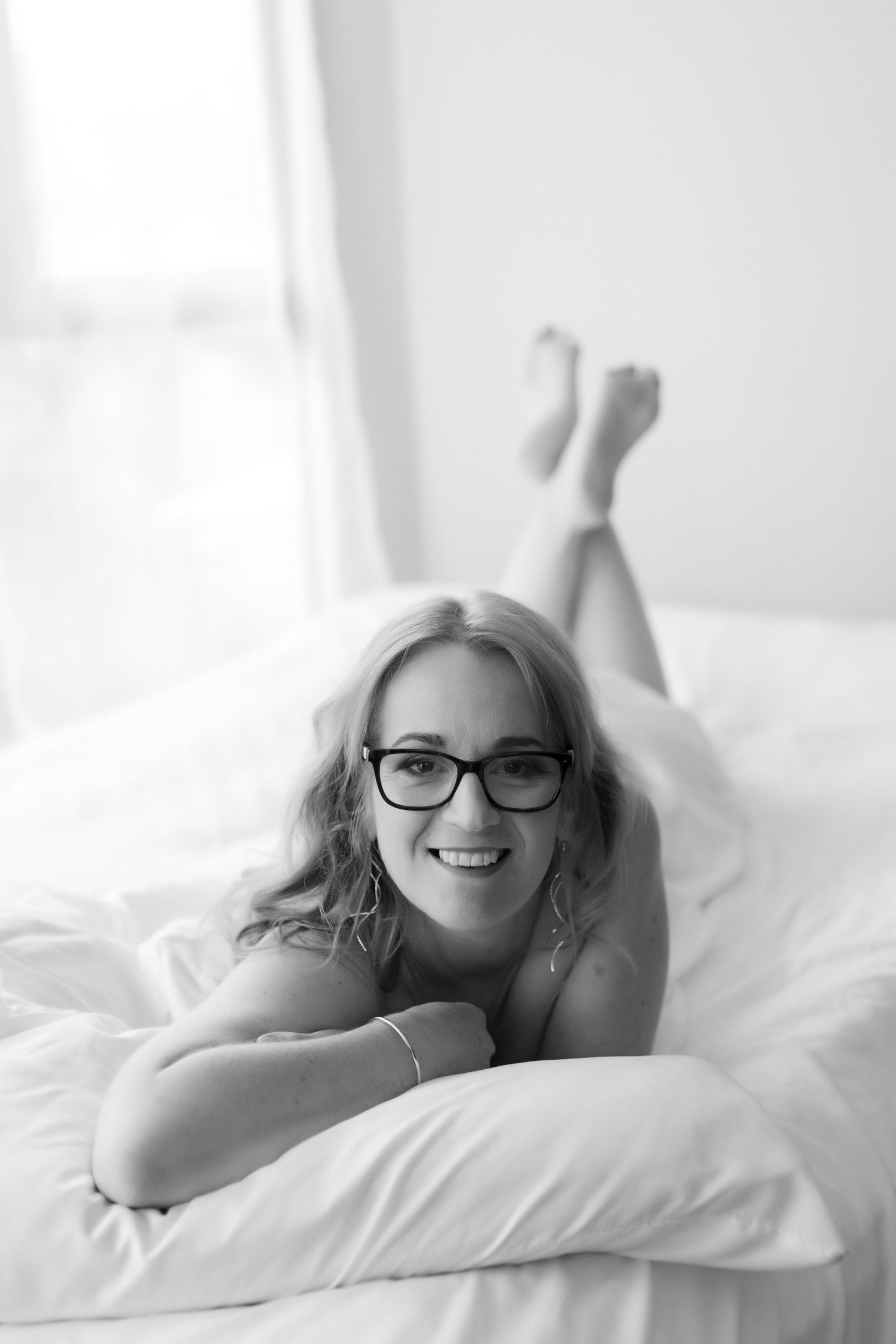 Smiling blonde woman wearing glasses lying on white bed, black and white boudoir photography. Boudoir photography Christchurch