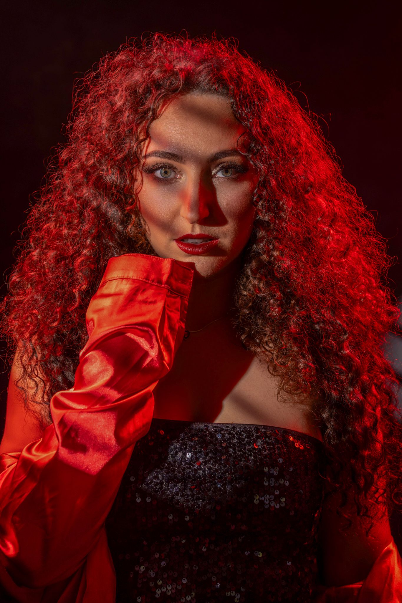Dramatic portrait of a curly-haired woman in red satin gloves under intense red lighting. Boudoir photography Christchurch portrait