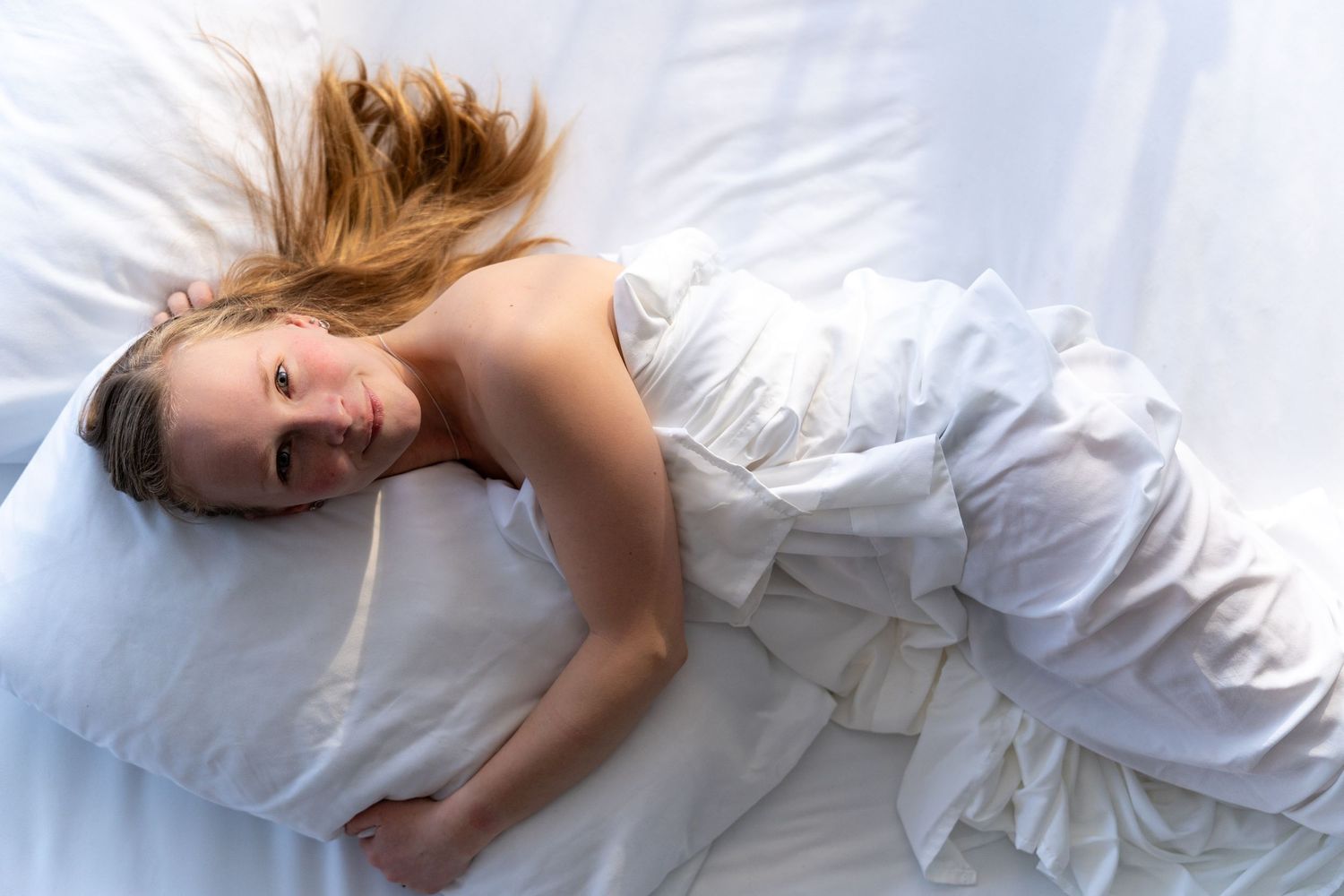 Woman with blonde hair lying in white bed covered with white sheets, looking relaxed and peaceful.