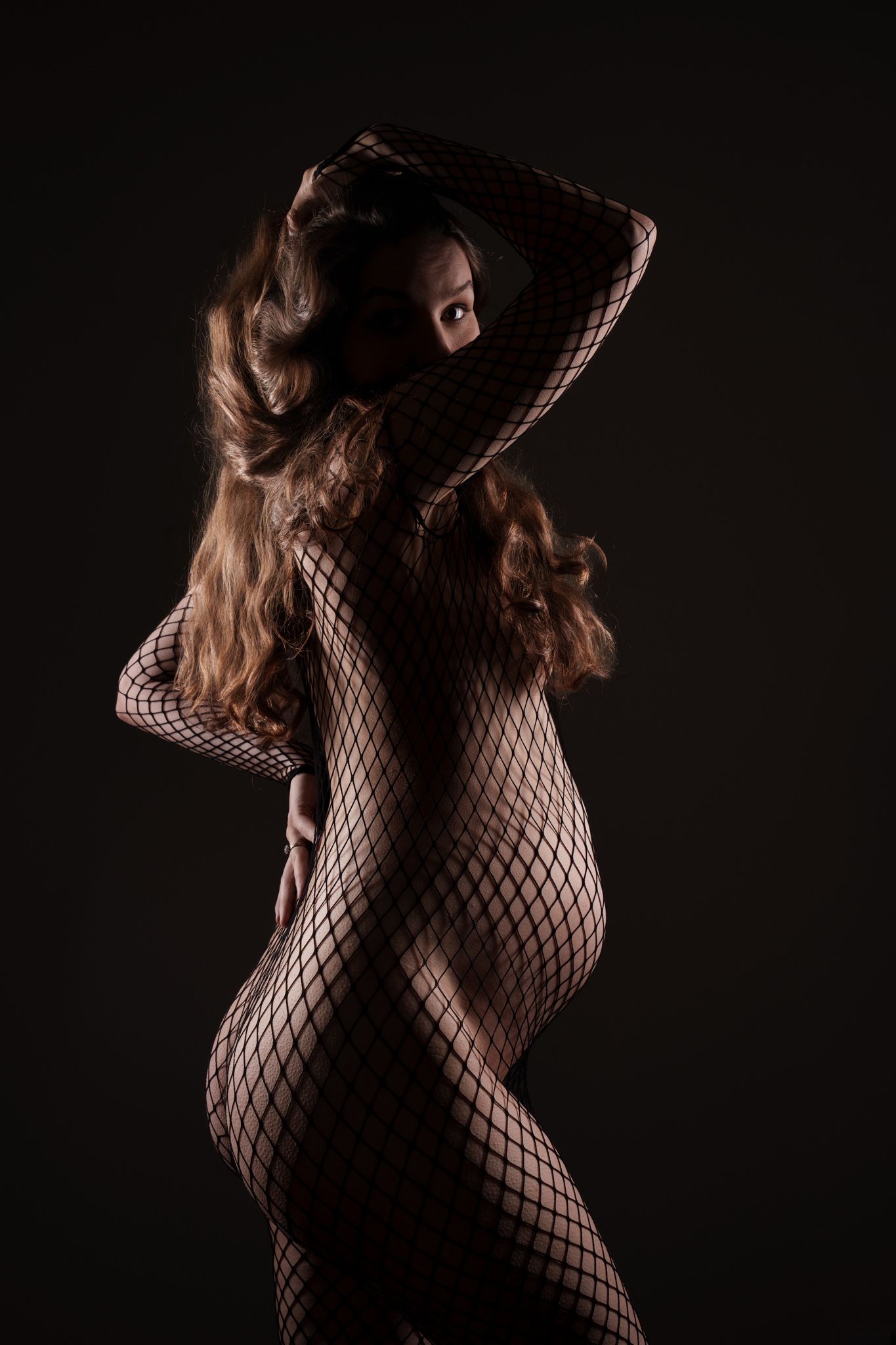 Silhouette of a pregnant woman in a fishnet bodysuit posing in dramatic studio lighting. Boudoir photography Christchurch maternity