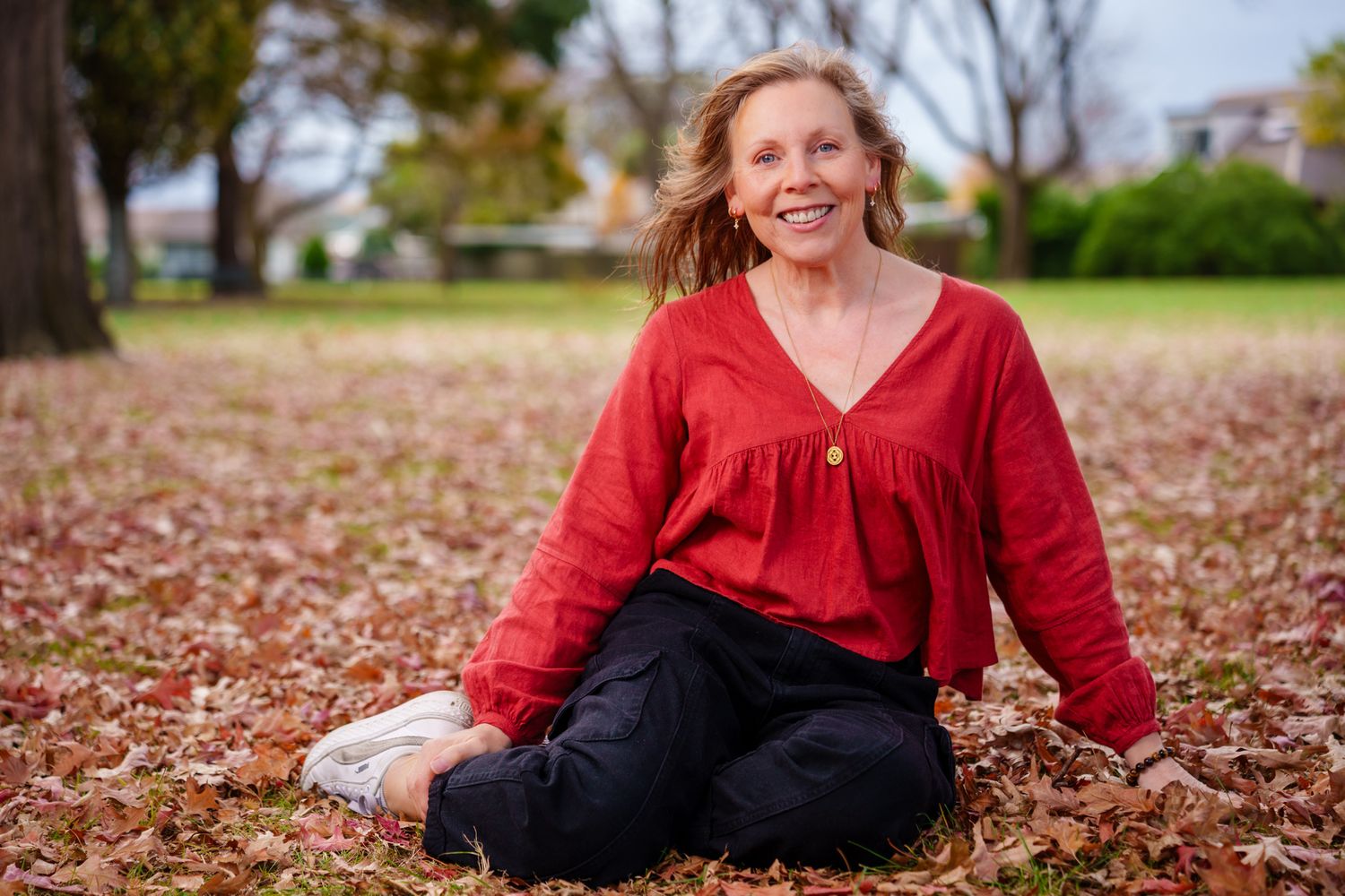 Smiling woman in red blouse sitting cross-legged on autumn leaves in a park setting. portrait photography Christchurch