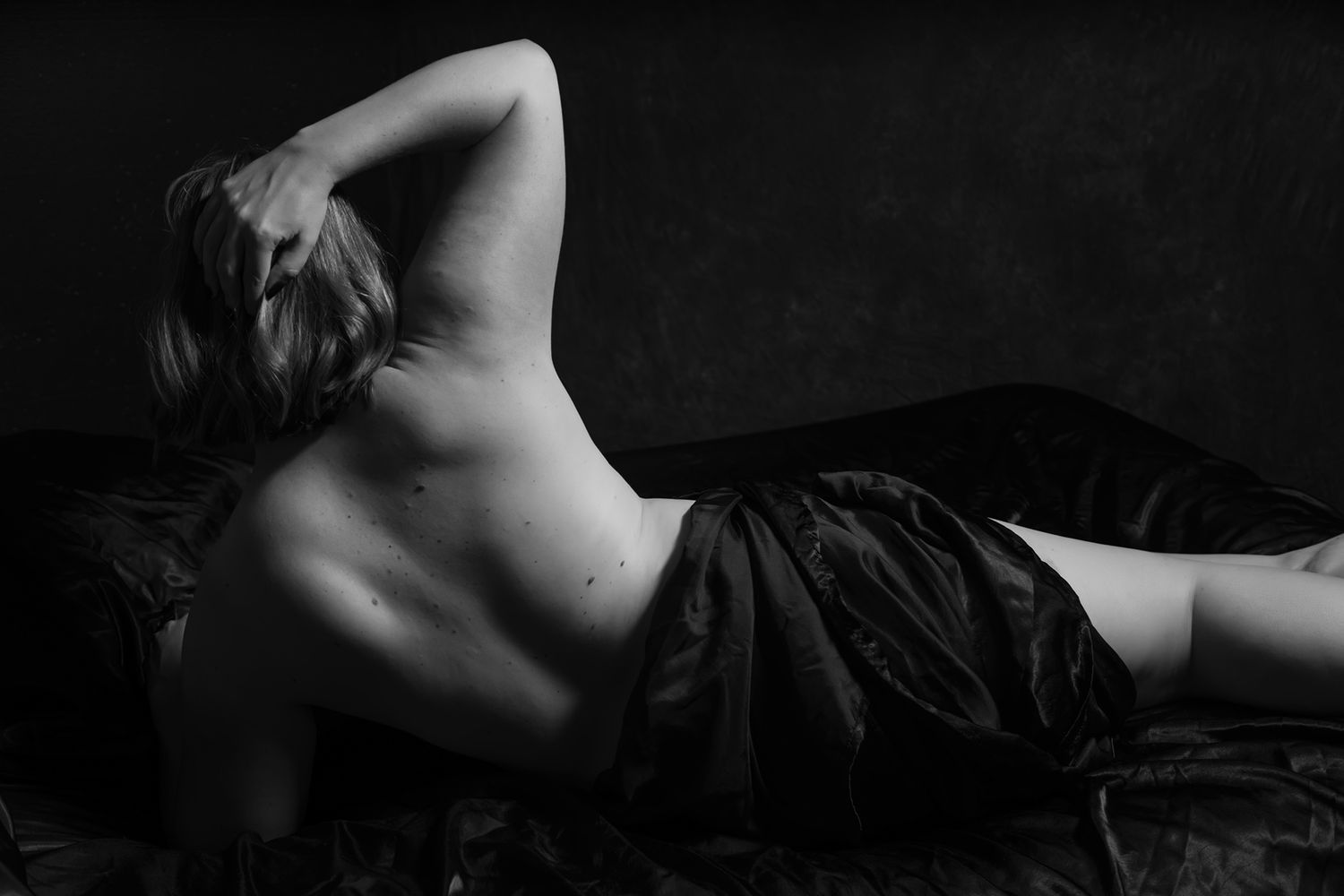 Black and white artistic photo of woman lying on dark satin sheets with back exposed. Boudoir photography Christchurch
