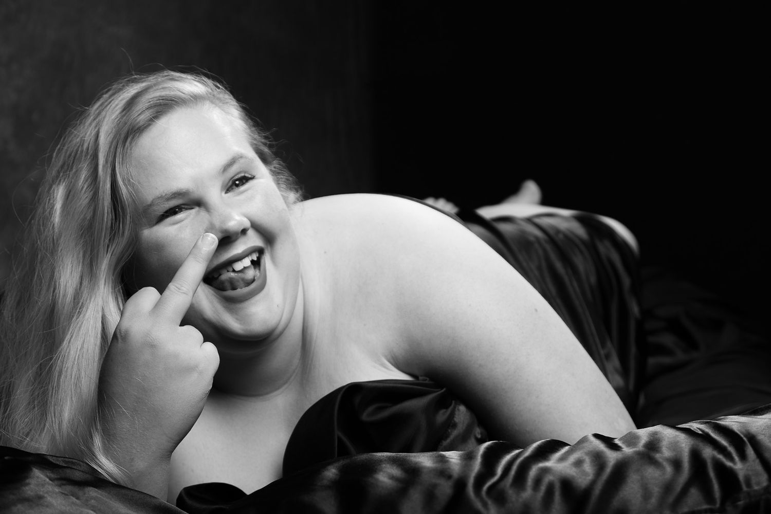 Black and white boudoir photo of a plus-size blonde woman laughing and posing playfully on a dark satin surface. Boudoir photography Christc