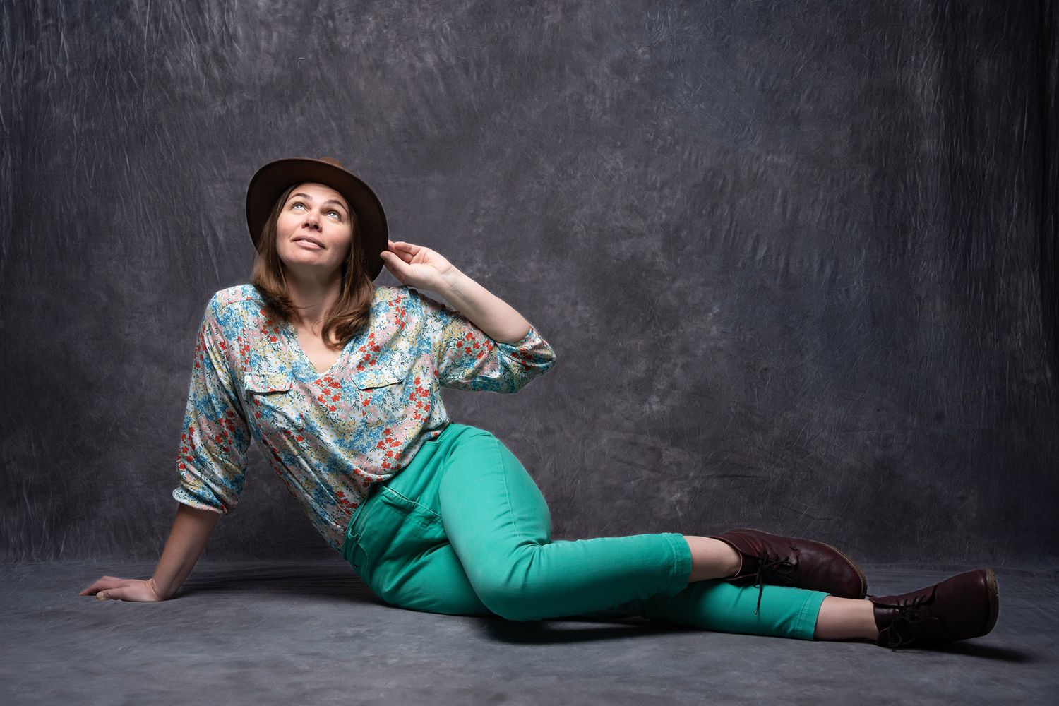 Stylish woman in floral blouse, green pants, and brown hat sitting on floor in a dark studio. portrait photography Christchurch