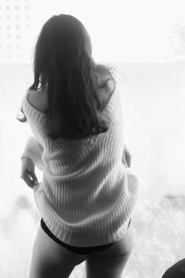 Black and white rear view of a woman in an oversized knit sweater and underwear by a bright window. Boudoir photography Christchurch