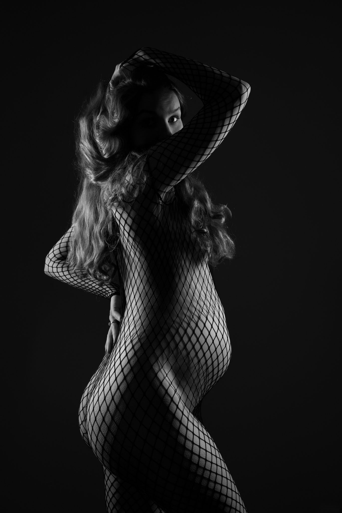 Black and white artistic photo of a woman in fishnet bodysuit posing gracefully against dark background.