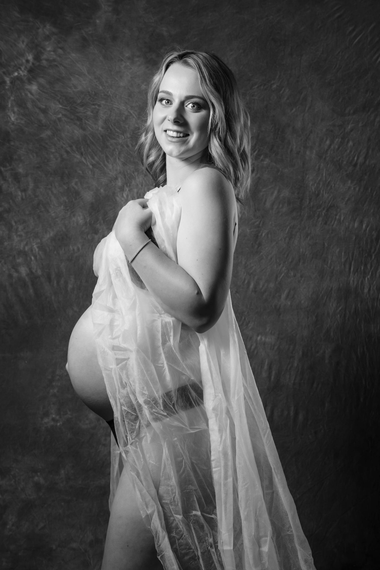 Smiling pregnant blonde woman wrapped in sheer white fabric poses in black and white maternity photo.  Boudoir photography Christchurch