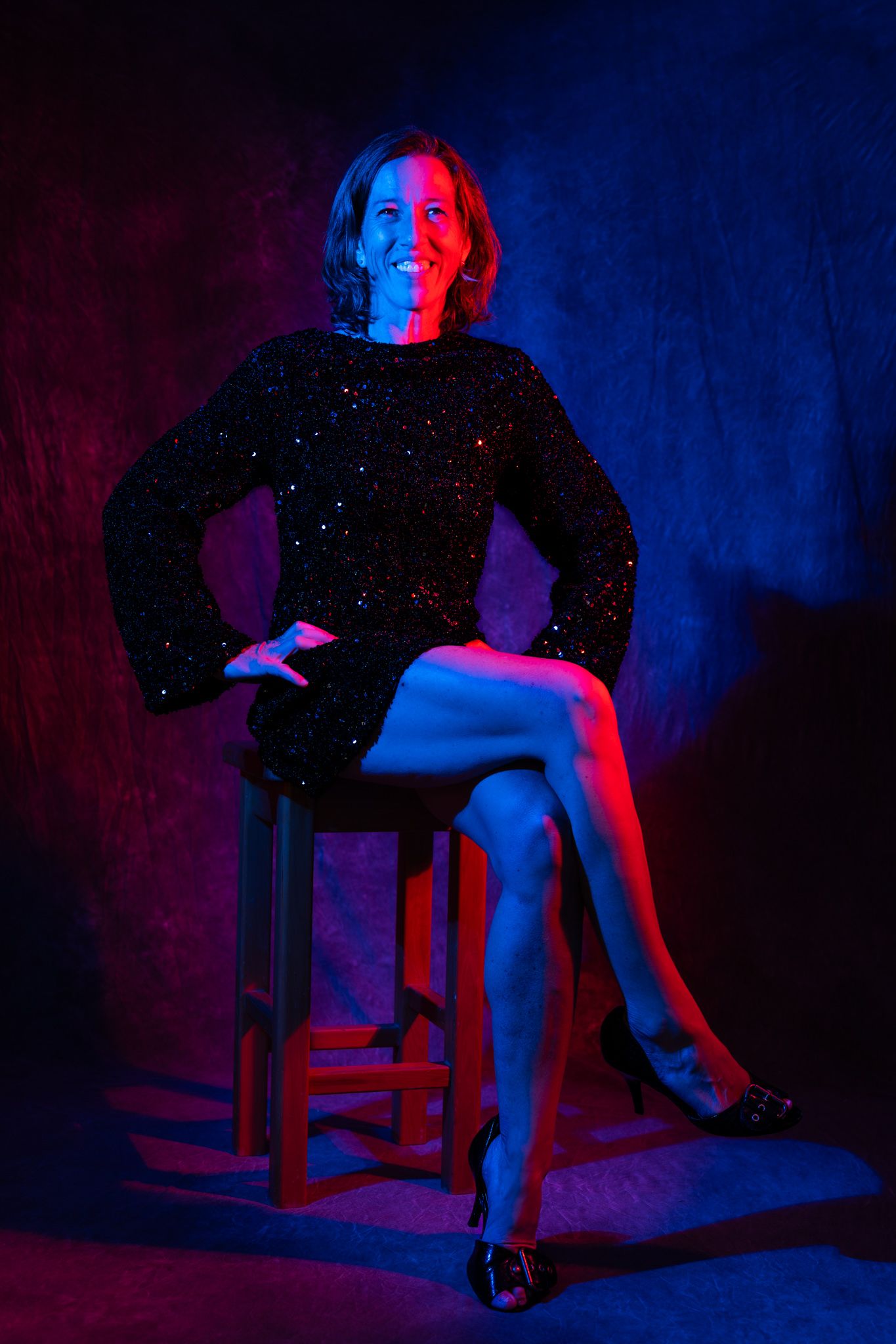 Woman in black sequin dress seated on stool under dramatic red and blue studio lighting, smiling confidently. Boudoir photography portrait