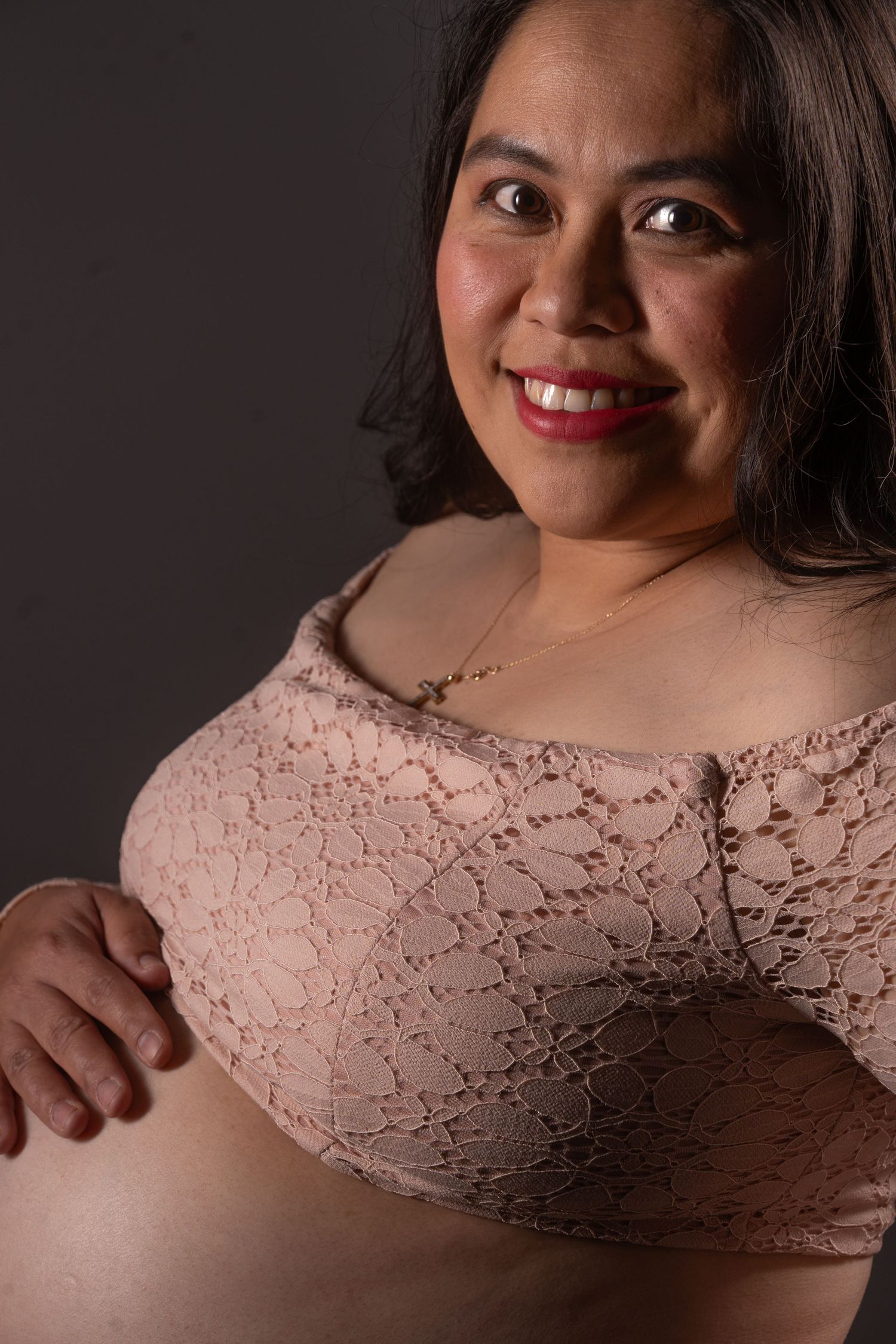 Smiling pregnant woman in pink lace bralette poses for maternity portrait on gray background. Boudoir photography Christchurch maternity