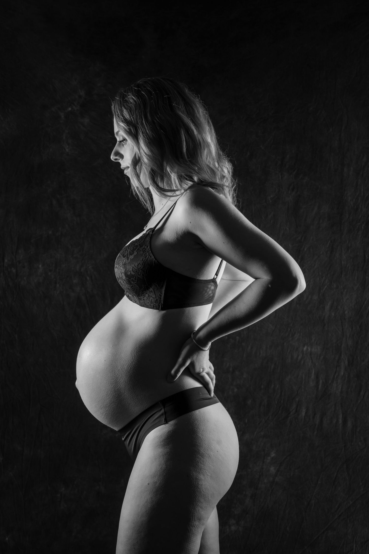 Black and white side profile of a pregnant woman in lingerie posing against a dark studio background.