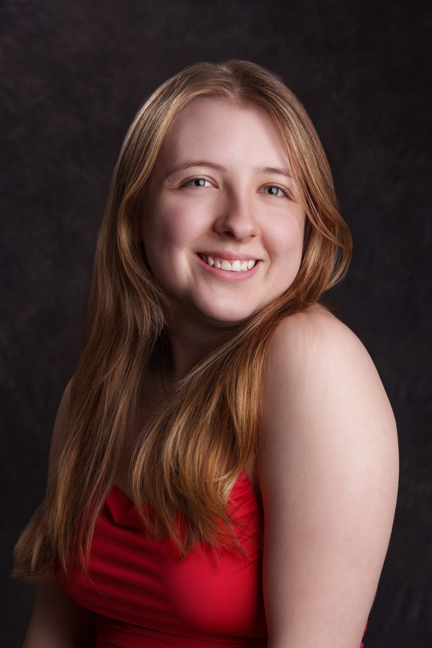 Smiling young woman with long auburn hair wearing a red dress against a dark studio backdrop. portrait photography Christchurch