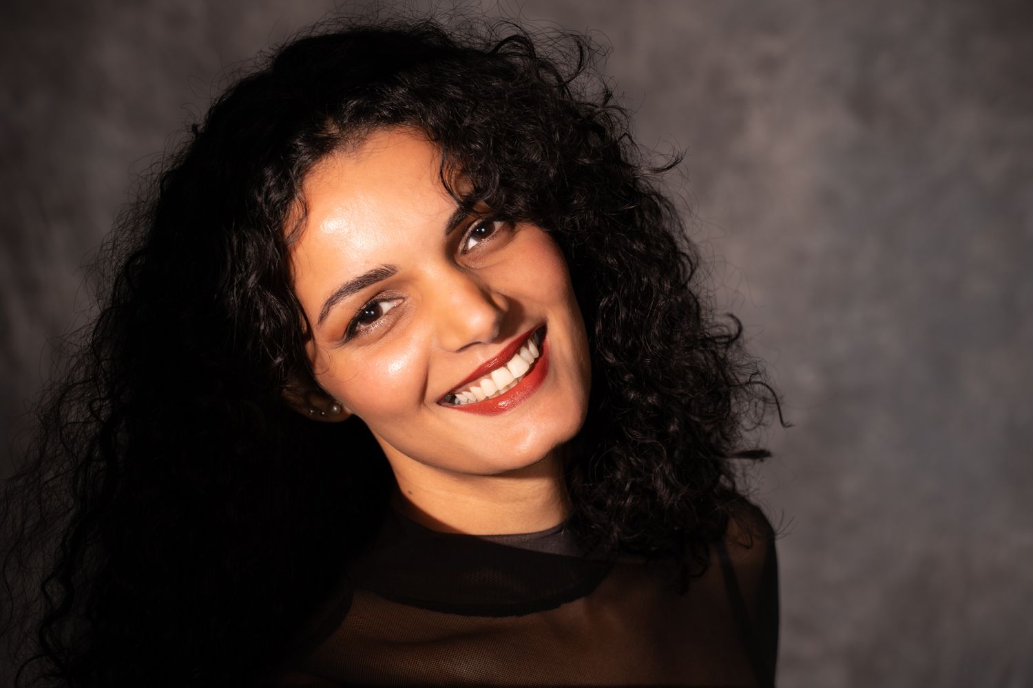 Smiling woman with long curly black hair wearing black mesh top against gray studio background. Boudoir photography Christchurch