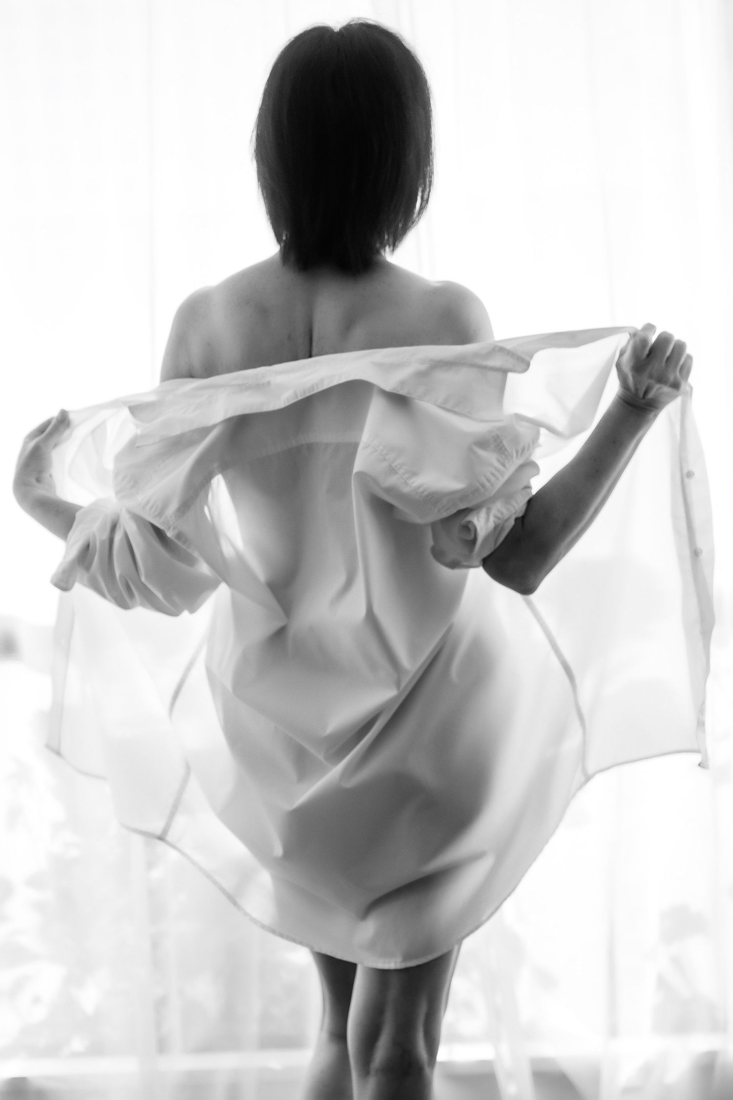 Black and white boudoir photo of a woman viewed from behind, holding a flowing white sheer fabric near a bright window. Boudoir photography