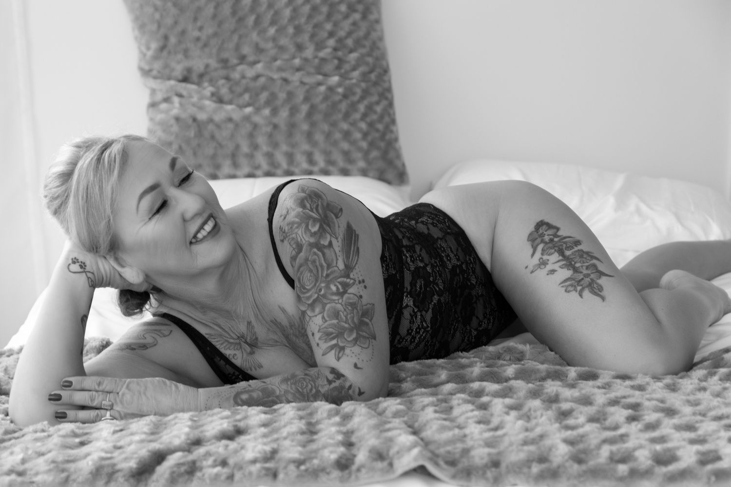 Black and white boudoir photo of a smiling tattooed blonde woman in lace lingerie lying on a bed. Boudoir photography Christchurch