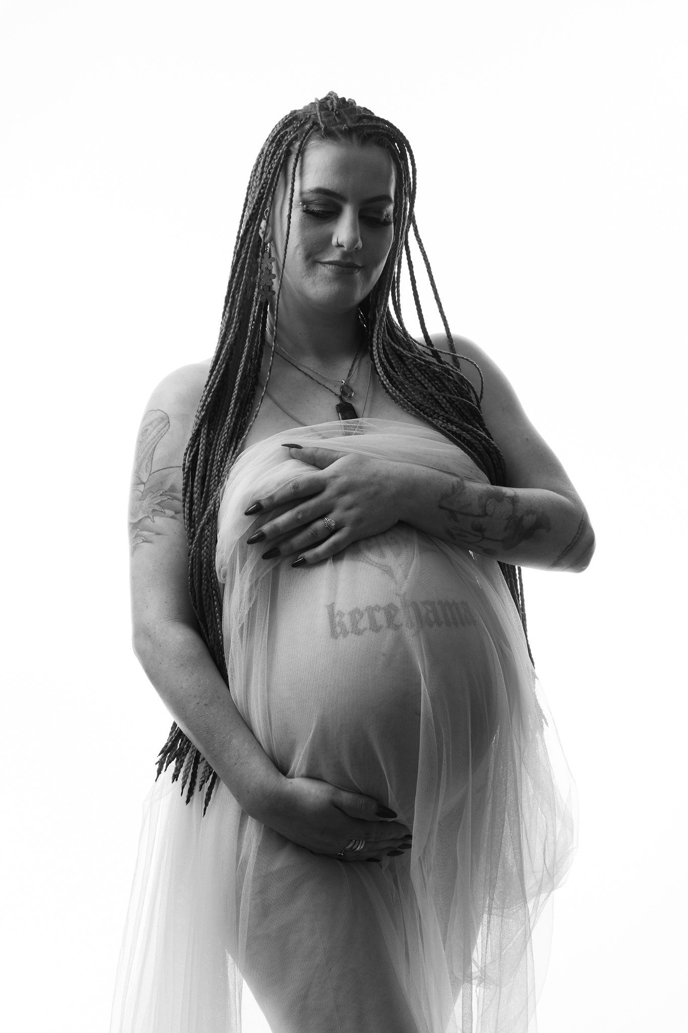 Black and white maternity photo of pregnant woman with braids cradling baby bump, wearing sheer fabric.