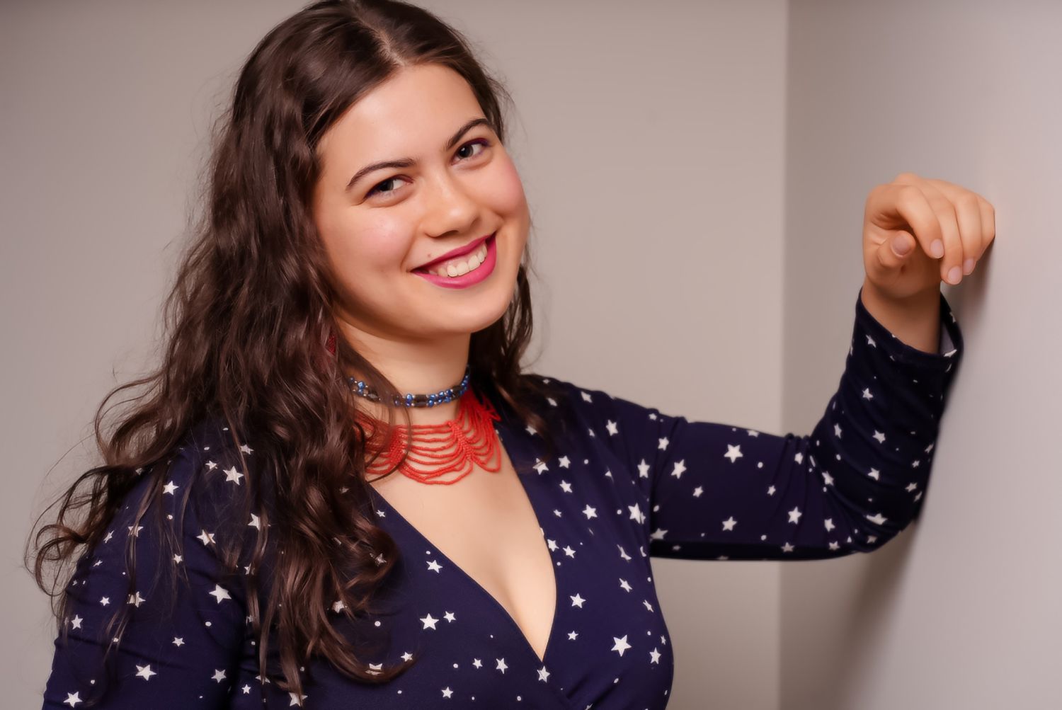 Cheerful woman in navy star-print dress with red choker necklace flexing arm against white wall. portrait photography Christchurch