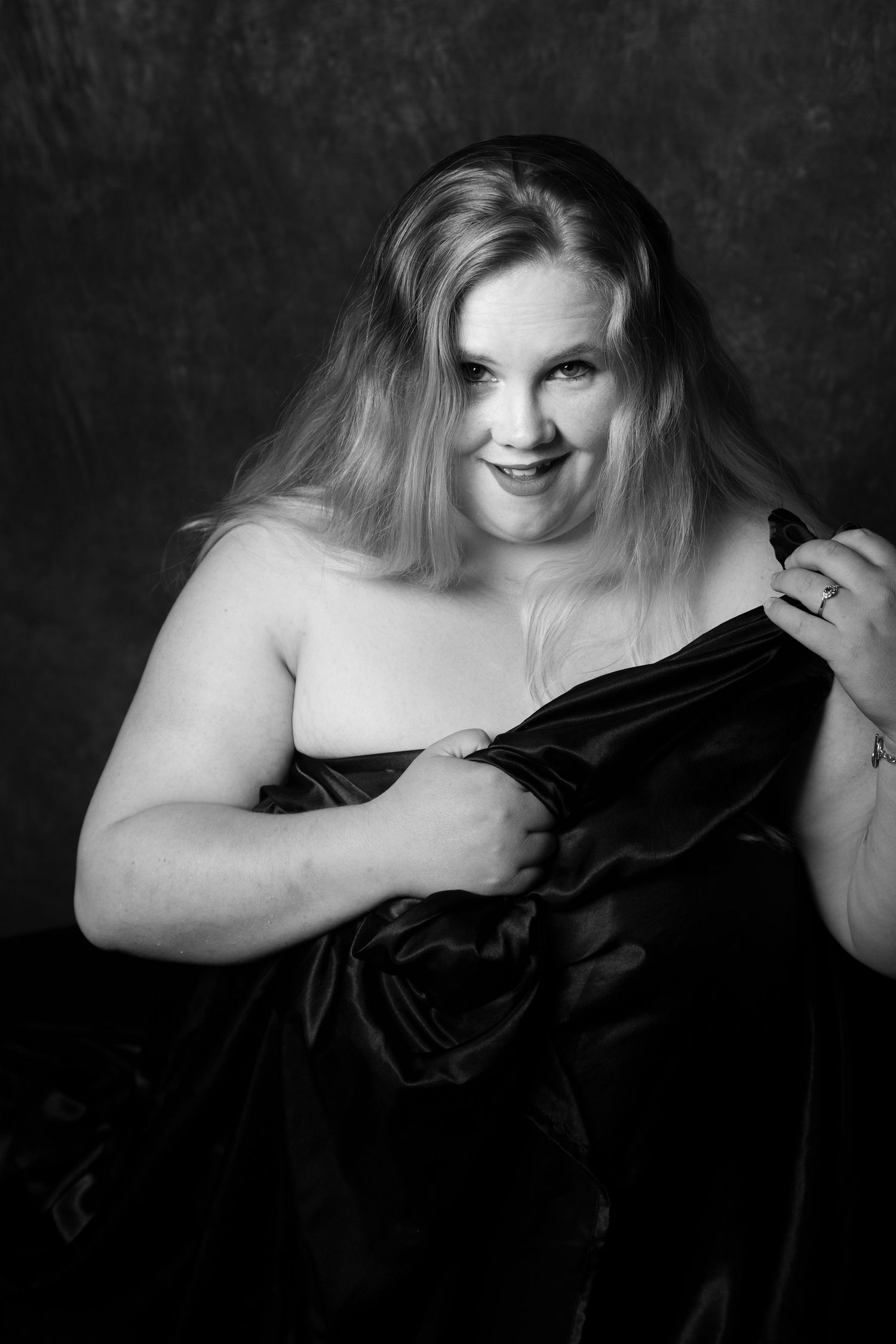 Black and white boudoir portrait of a plus-size blonde woman holding dark satin fabric while smiling confidently. Boudoir photography