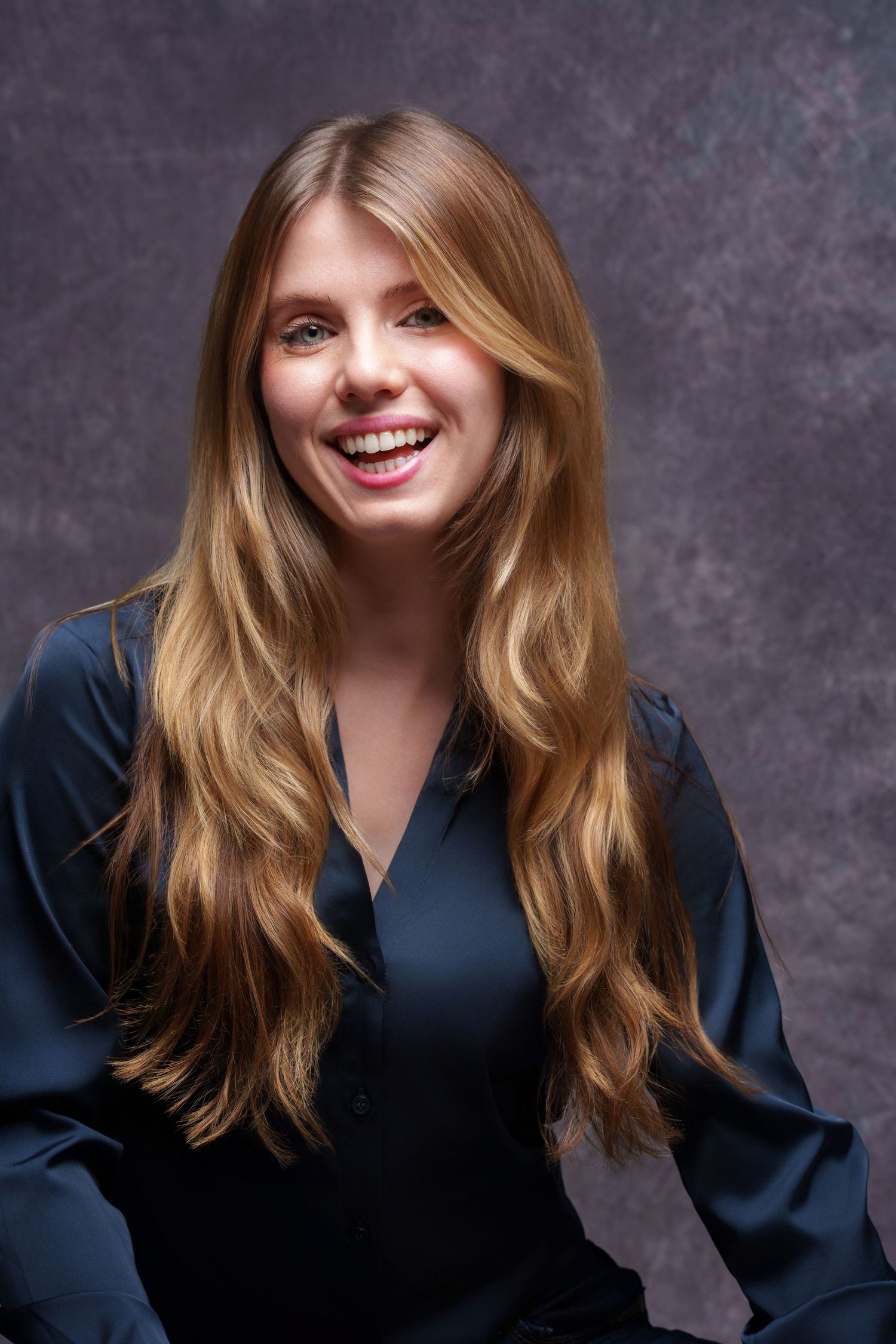 Cheerful blonde woman with long wavy hair laughing in navy blue blouse against gray backdrop. portrait photography Christchurch
