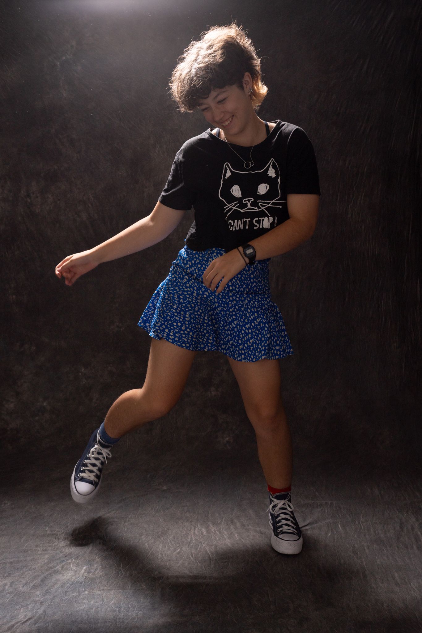 Young woman dancing in black cat tee and blue floral skirt with Converse sneakers against dark backdrop. portrait photography Christchurch