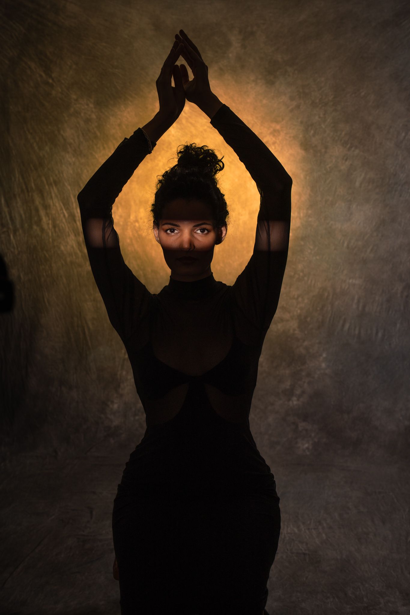 Silhouetted woman in black backless dress with hands raised against golden glowing background. Boudoir photography Christchurch