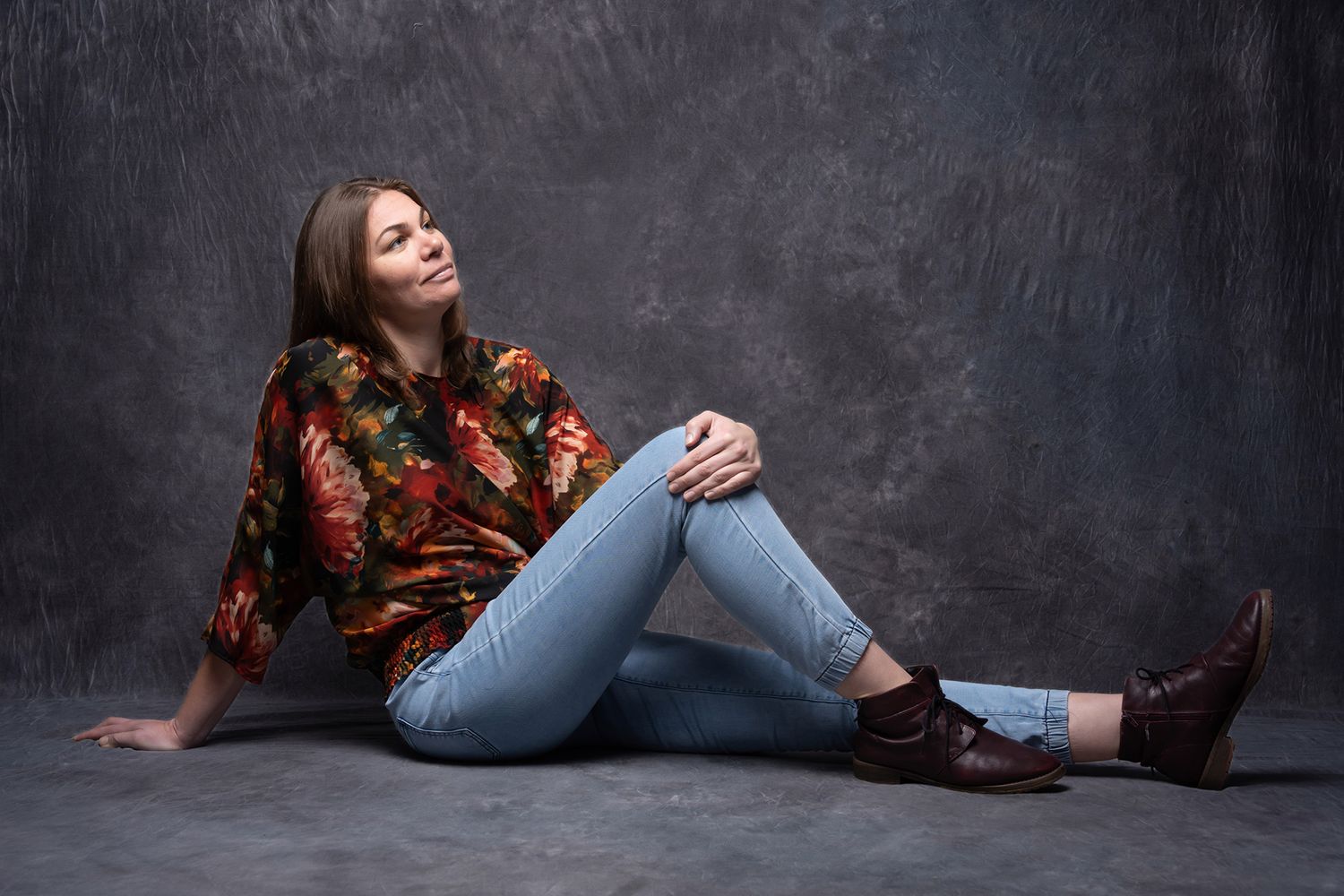 Young woman in floral blouse and jeans sitting on floor, smiling upward in a studio portrait. portrait photography Christchurch