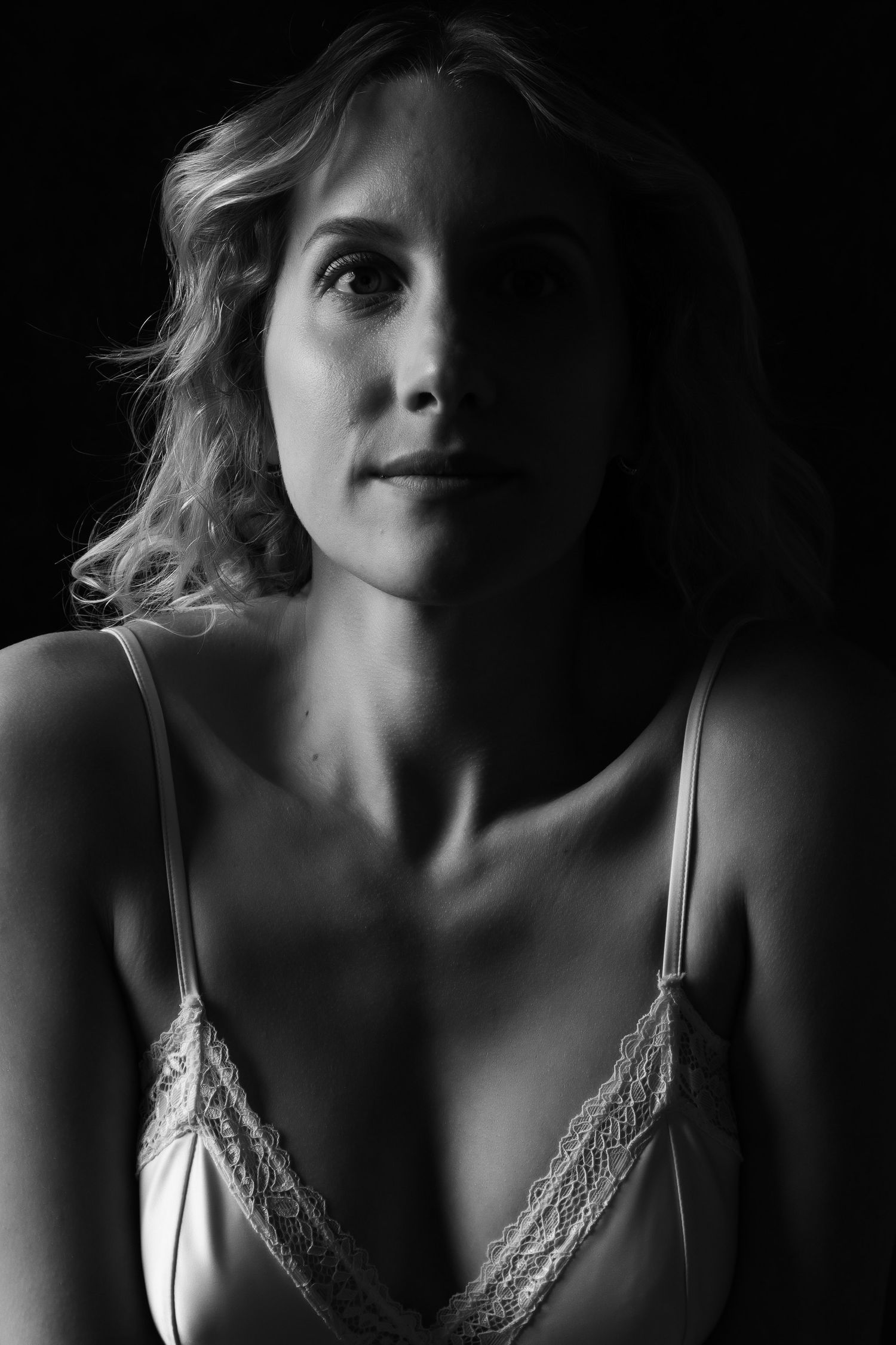 Black and white low-key portrait of a blonde woman in a lace bralette looking upward thoughtfully. Boudoir photography Christchurch