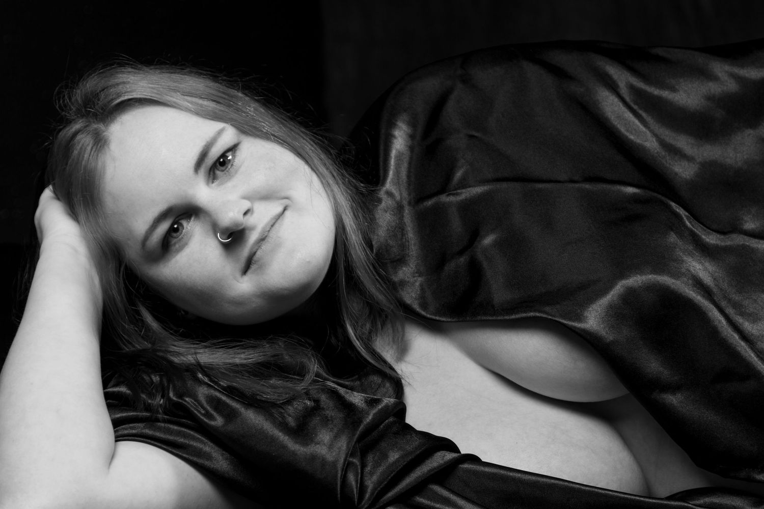 Black and white close-up portrait of a smiling woman wrapped in dark satin fabric lying down. Boudoir photography Christchurch