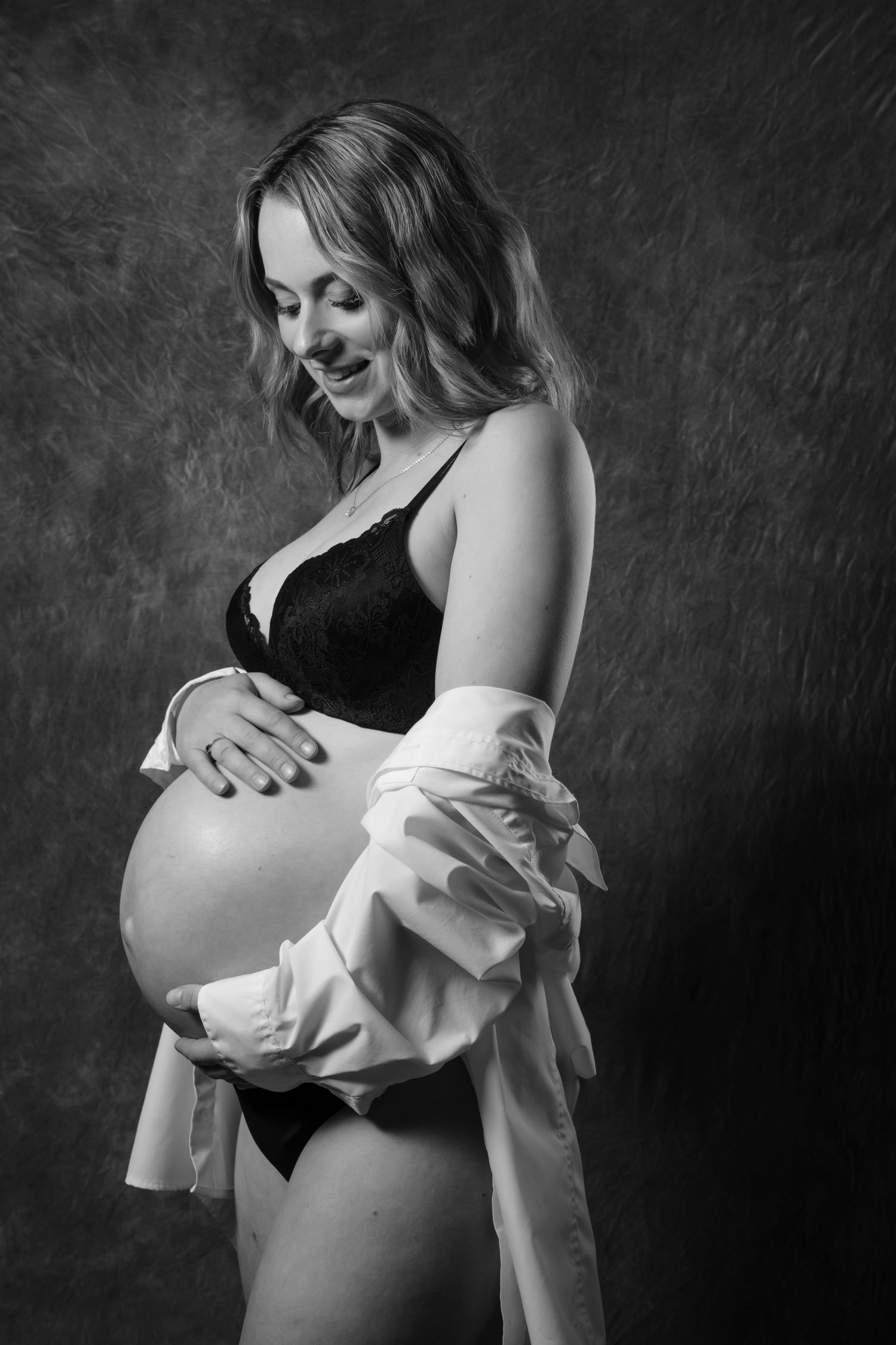 Black and white portrait of a smiling pregnant woman in lingerie and open white shirt cradling her baby bump.