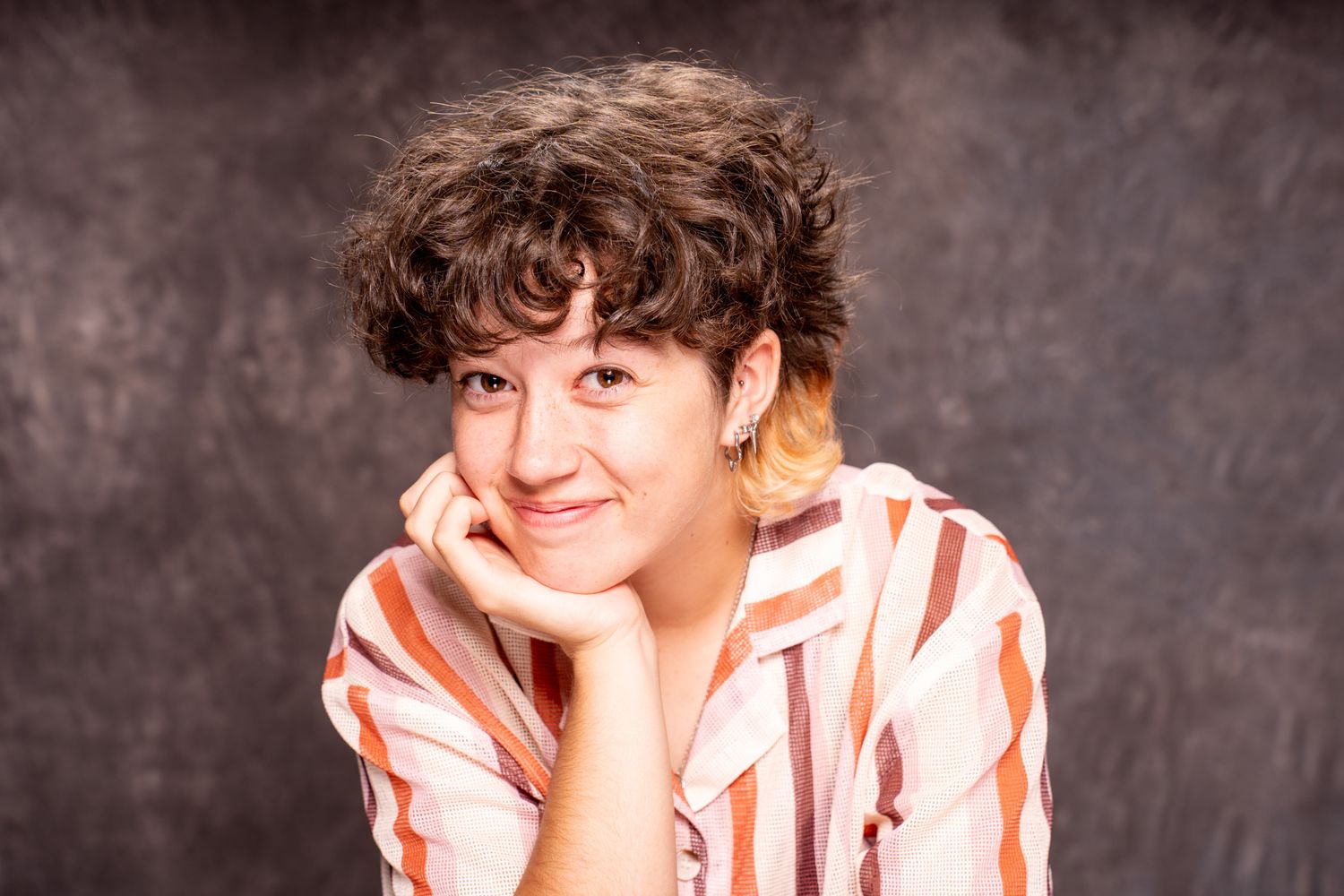 Cheerful person with short curly hair in a striped shirt, chin resting on hand, against a dark studio background. portrai photography Christ