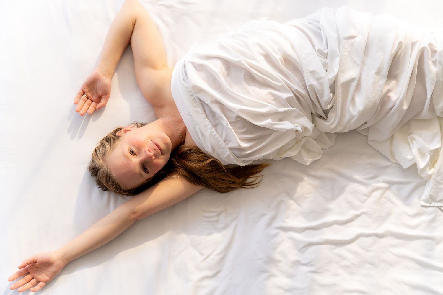 Young woman lying on white bedding wrapped in a white sheet, arms outstretched, viewed from above.