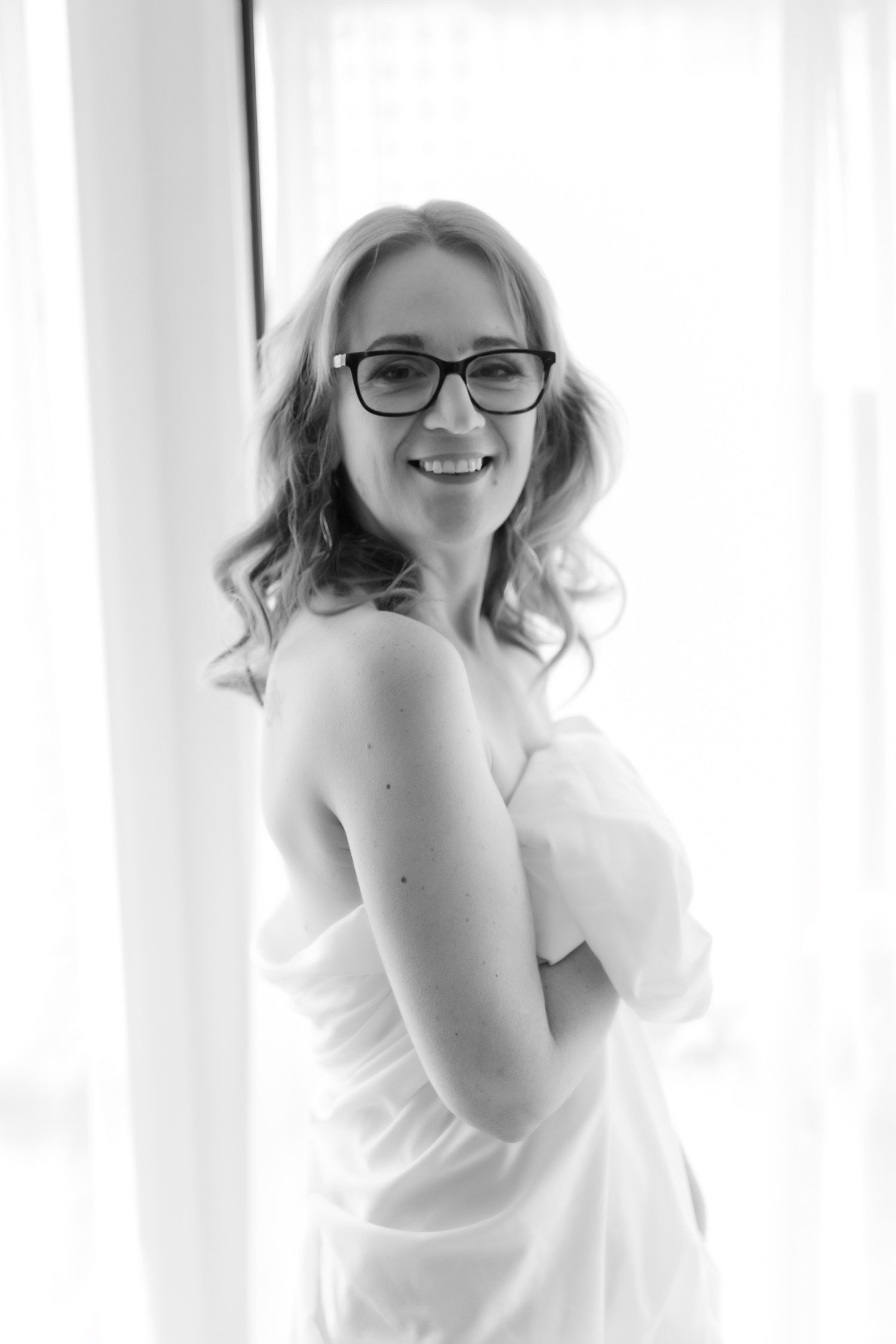 Smiling woman in glasses wrapped in white towel standing by window, black and white portrait. Boudoir photography Christchurch
