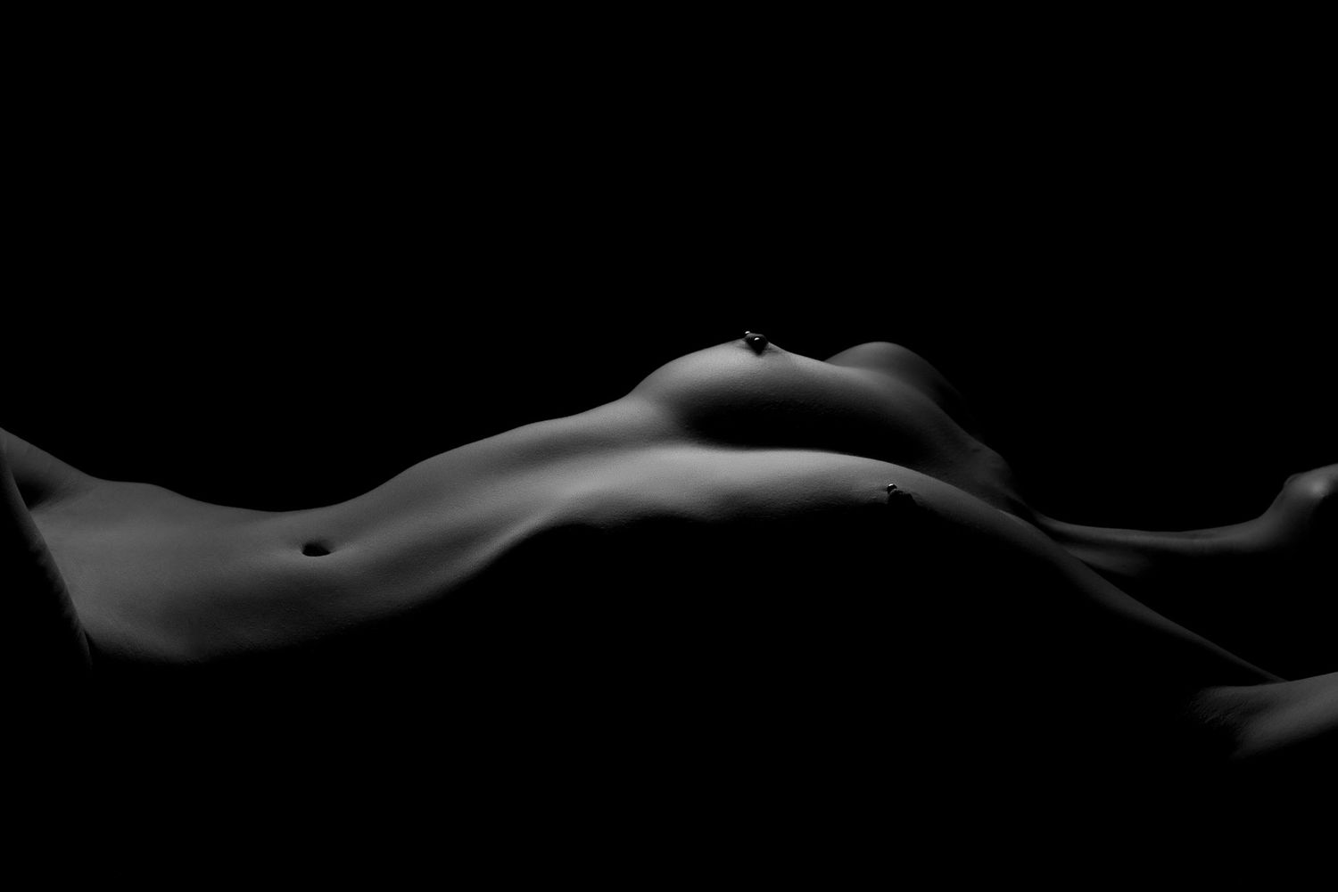 Black and white artistic bodyscape nude photograph with dramatic lighting showing feminine curves on dark background. Boudoir photography