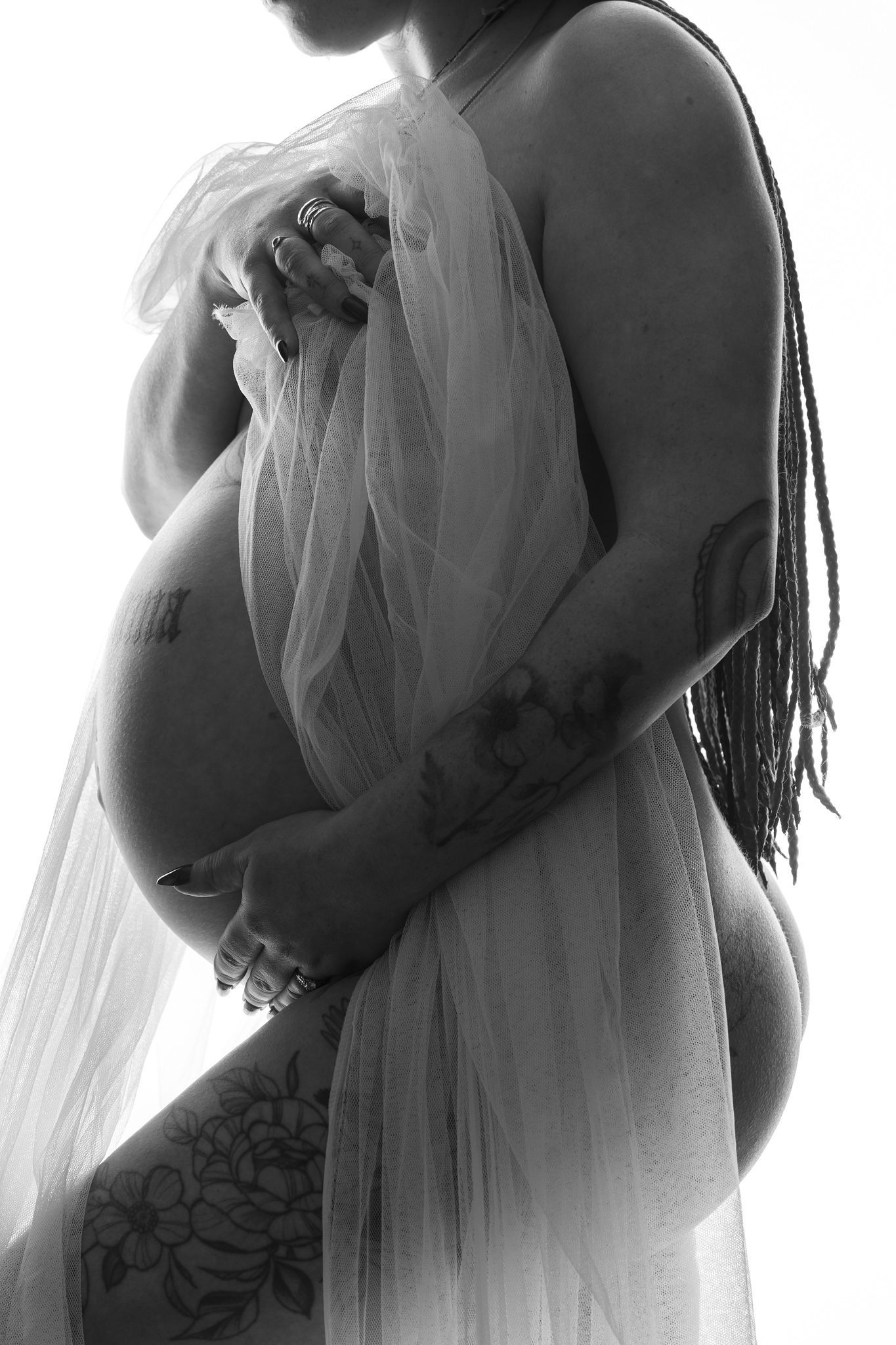 Black and white maternity photo of tattooed pregnant woman draped in sheer fabric, cradling her baby bump.