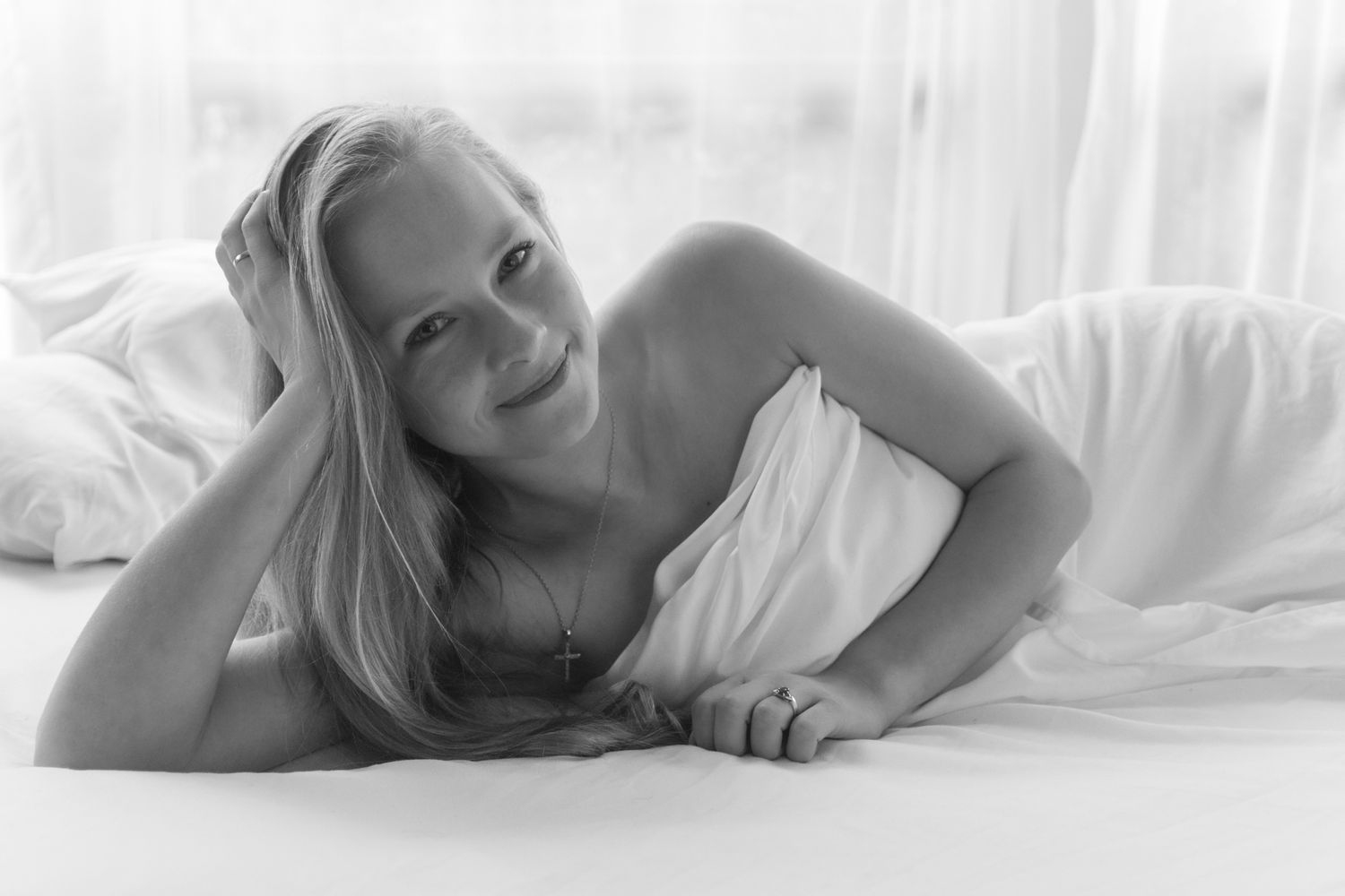 Soft black and white boudoir photo of a smiling blonde woman lying on white bedding wrapped in a sheet. Boudoir photography Christchurch