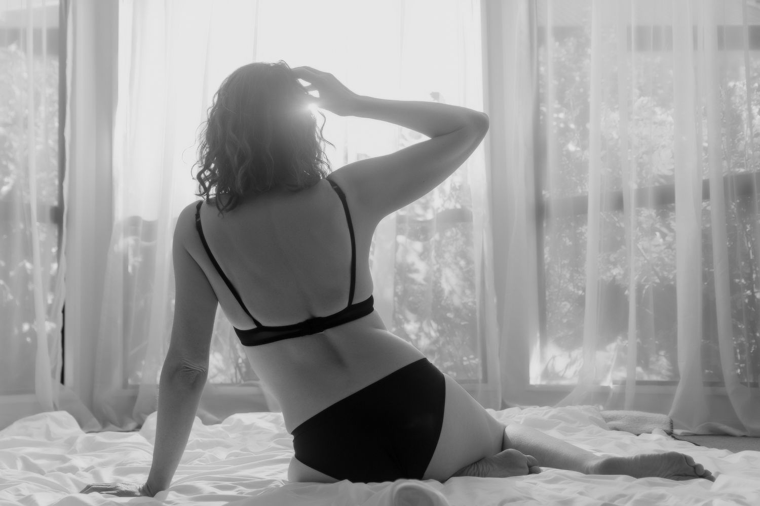 Black and white boudoir photo of a curvy woman in lingerie sitting on a bed facing sheer curtained windows. Boudoir photography Christchurch