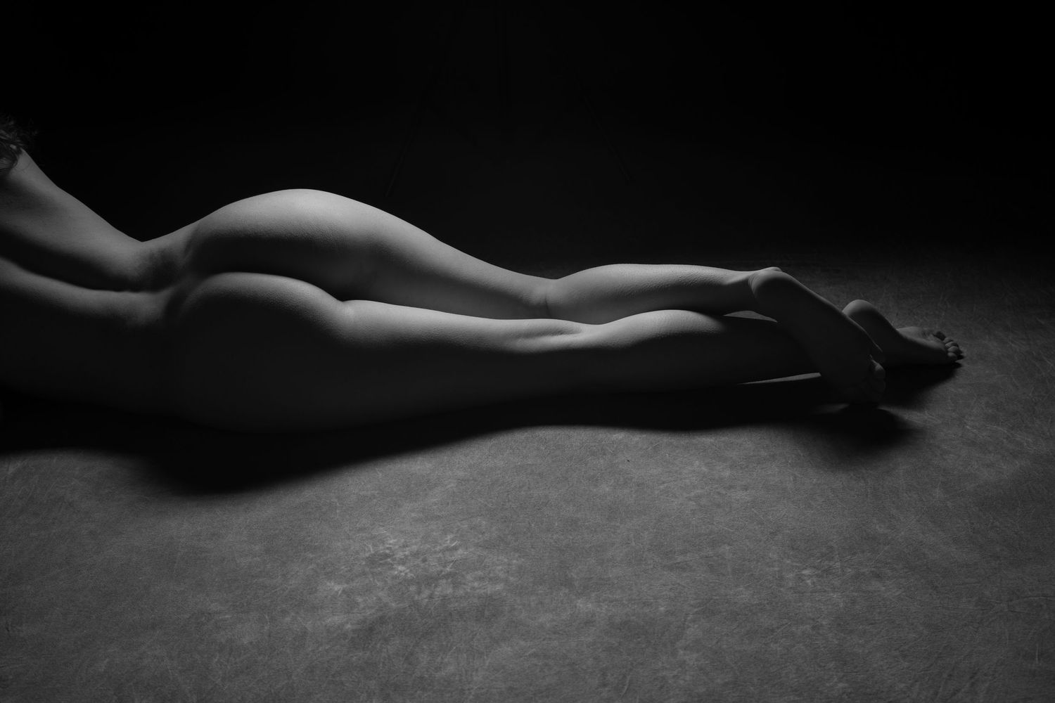 Black and white artistic bodyscape of a reclining nude figure with legs extended on a dark floor. Boudoir photography Christchurch